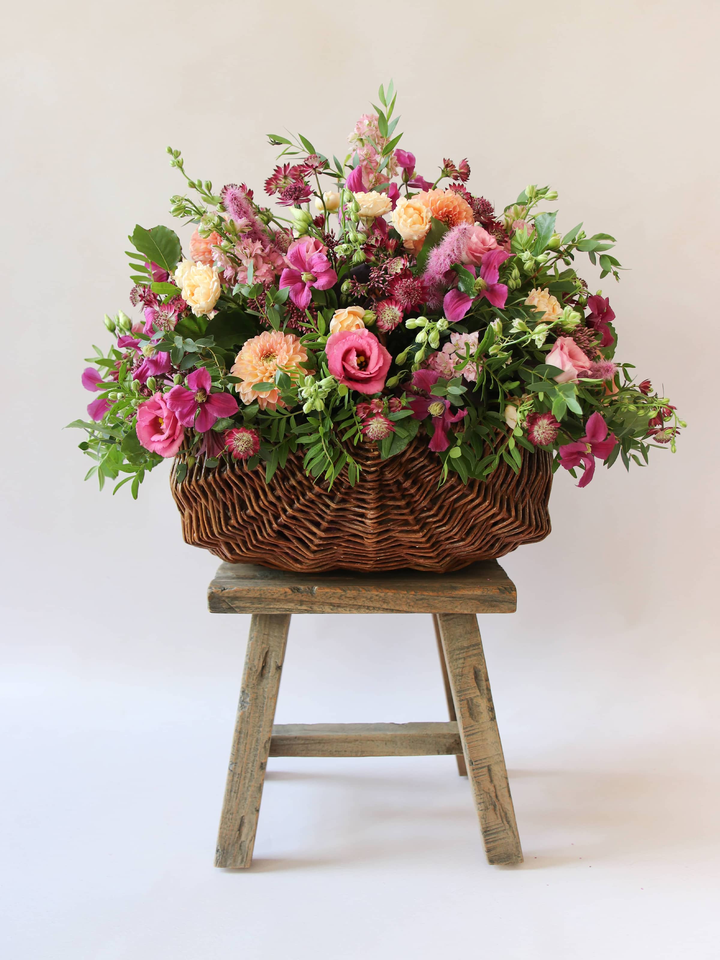 Arrangement of bright pink and peach flowers with green foliage in a wicker basket on a wooden stool against a light background