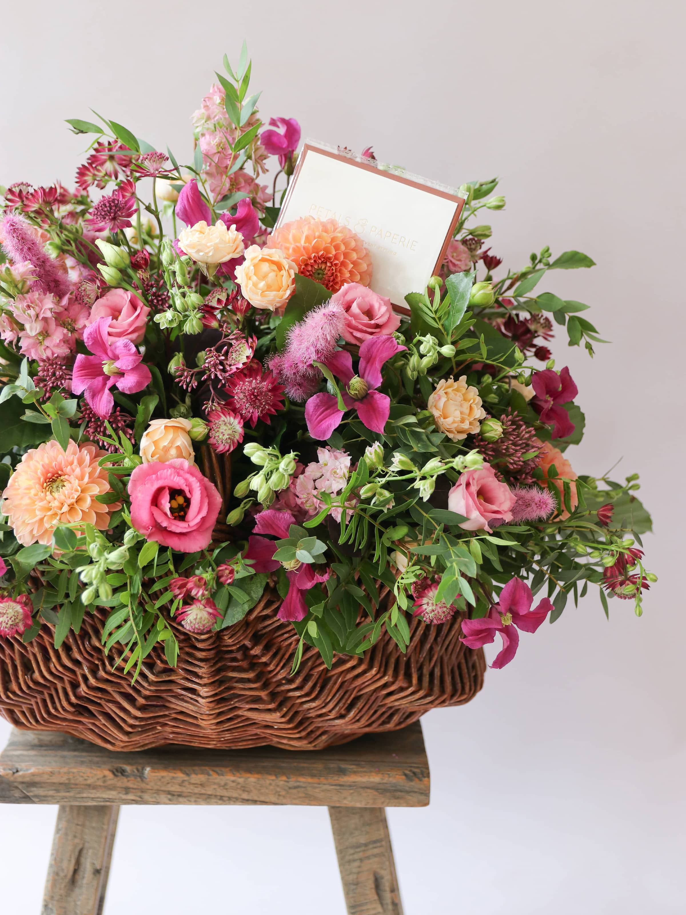 Arrangement of bright pink and peach flowers with green foliage in a wicker basket on a wooden stool against a light background with a Petals & Paperie Message card tucked inside the arrangement.