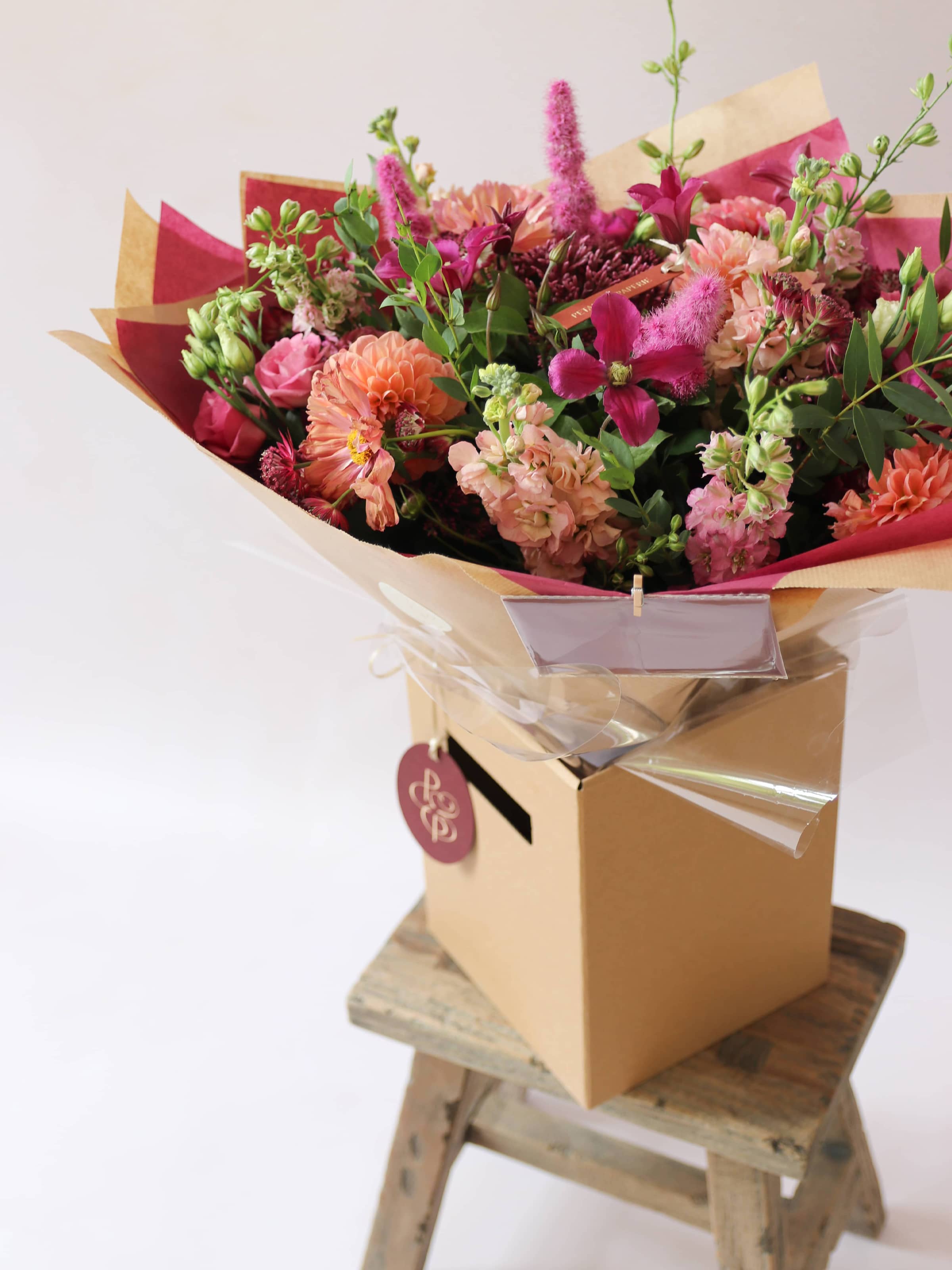 Bouquet of bright pink and peach flowers wrapped in brown paper on a wooden stool with a white background