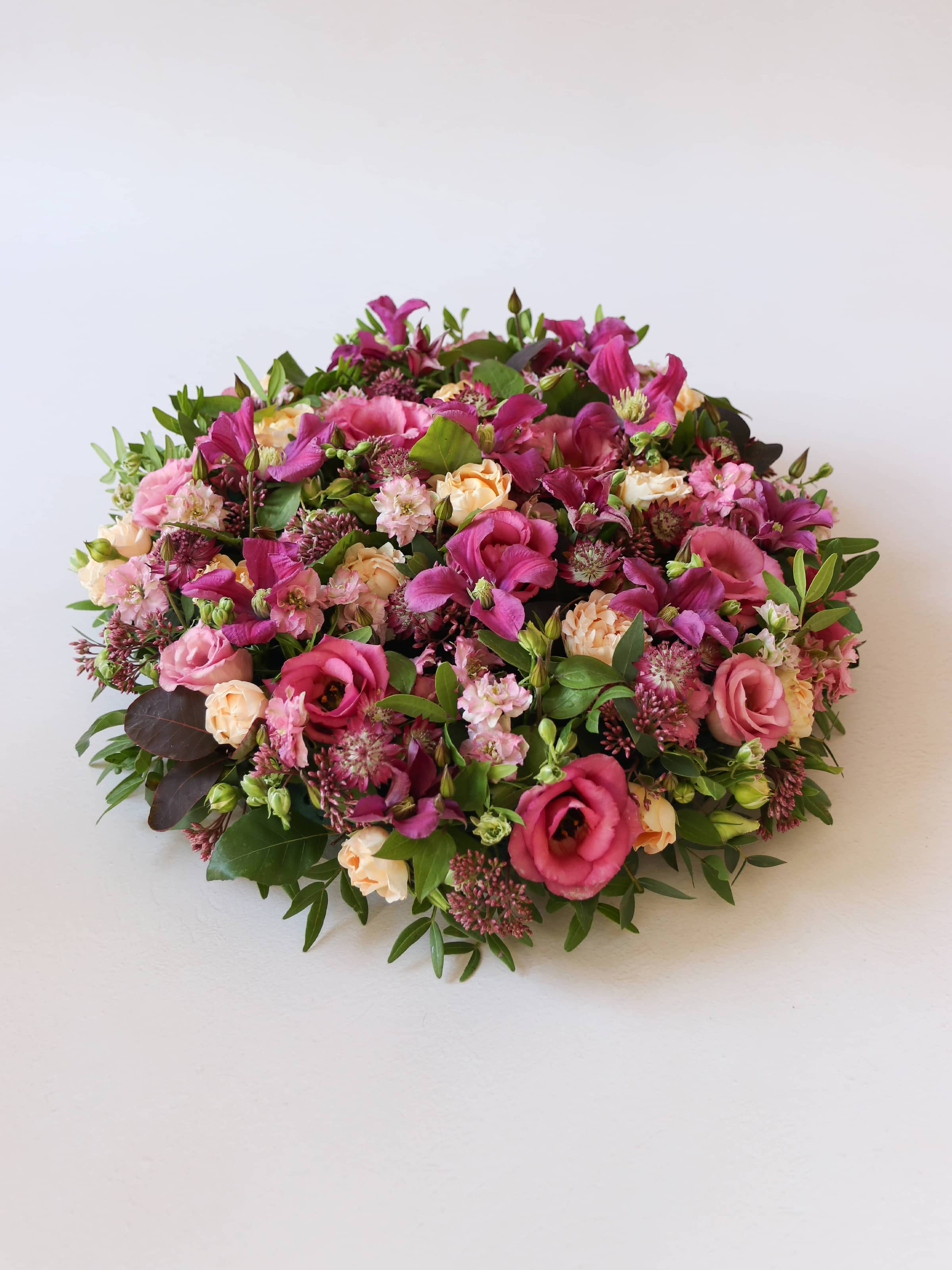 Angled view of a round funeral posy of bright pink and peach flowers with green foliage on a white background