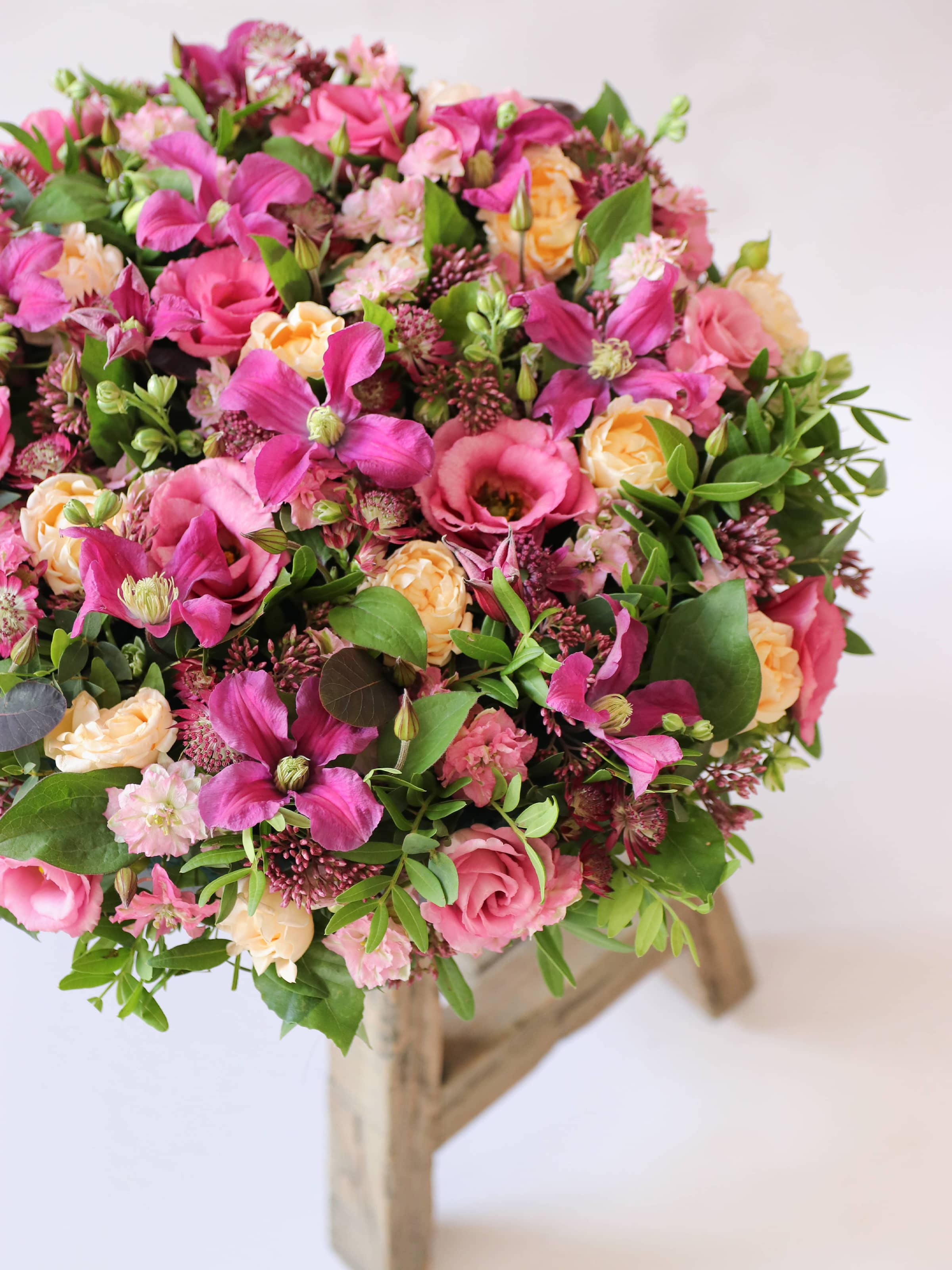 Angled view of a round funeral posy of bright pink and peach flowers with green foliage atop a wooden stool on a white background.