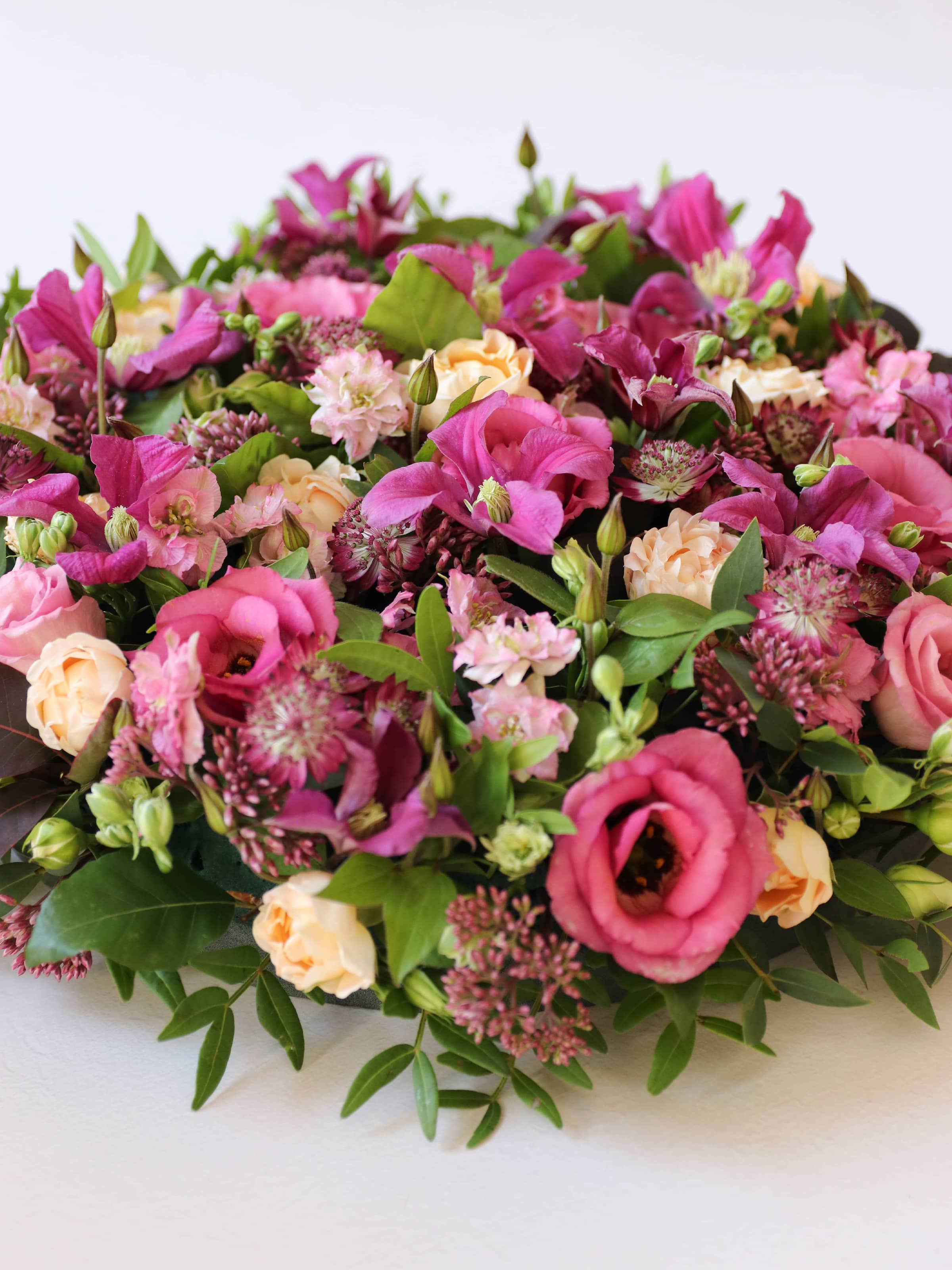 Close up view of a round funeral posy of bright pink and peach flowers with green foliage on a white background