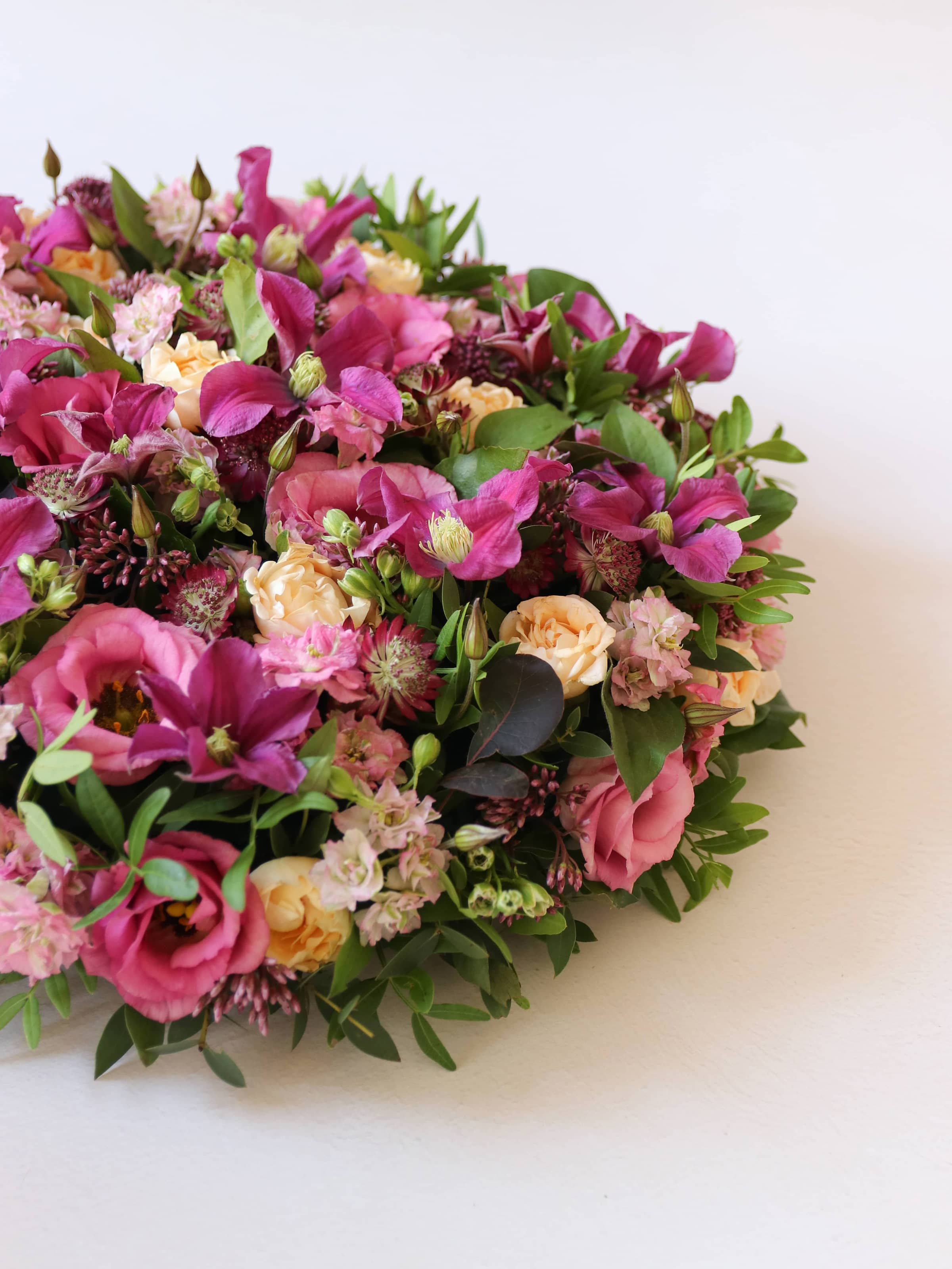 Top down side view of a round funeral posy of bright pink and peach flowers with green foliage on a white background