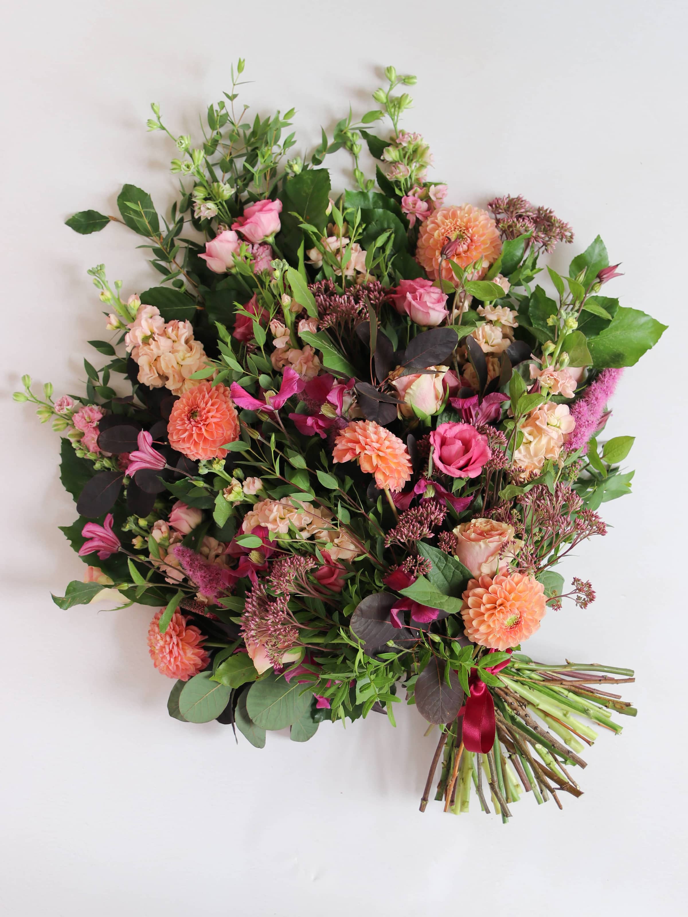 Portrait photograph of a tied funeral sheaf with flowers including pink Lisianthus, peach Stocks, pink Delphiniums and peach Dahlias, tied with a burgundy satin bow.