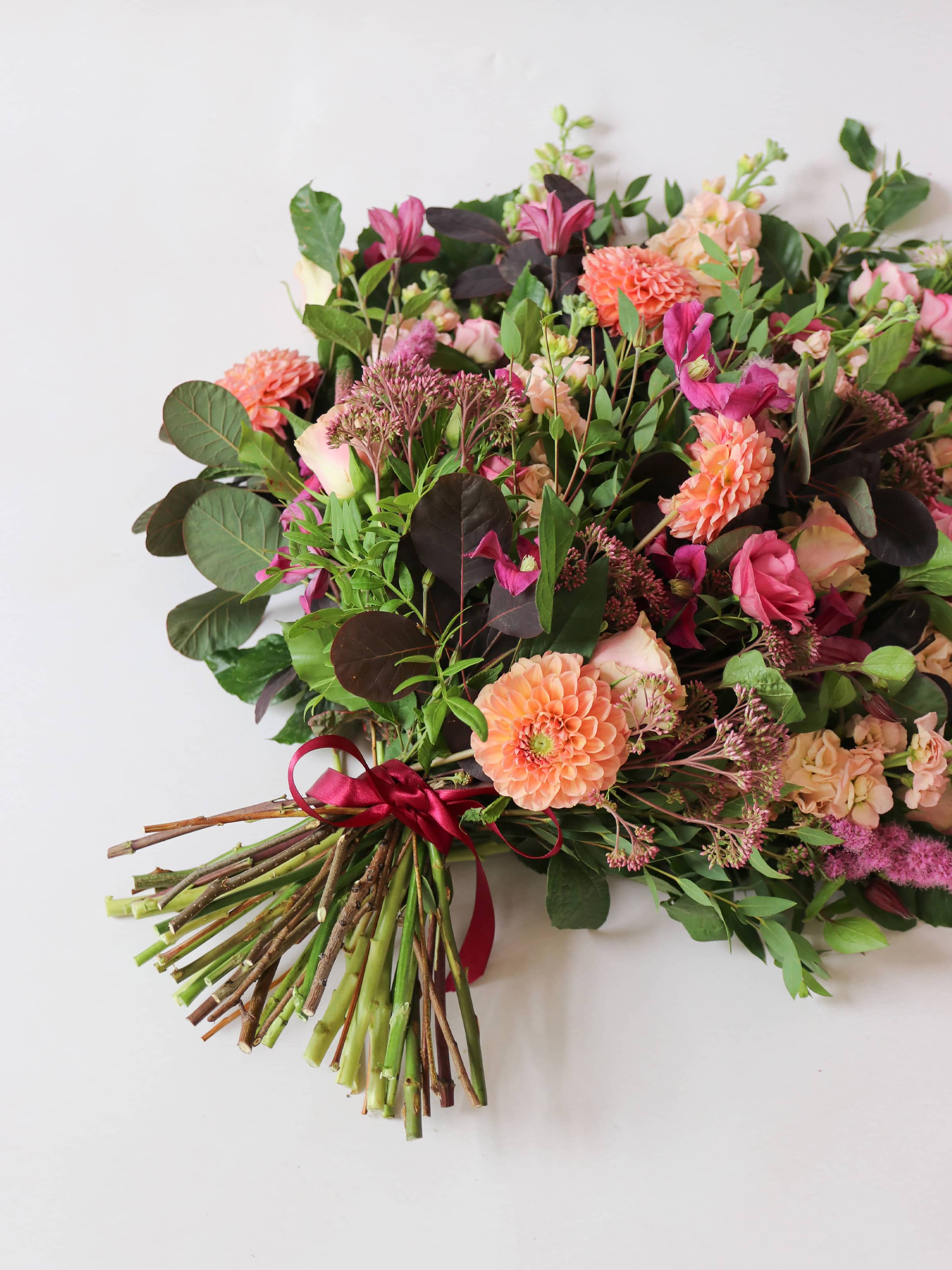 Colourful, top down view of a tied funeral sheaf with pink, peach and burgundy flowers, tied with a burgundy satin bow.