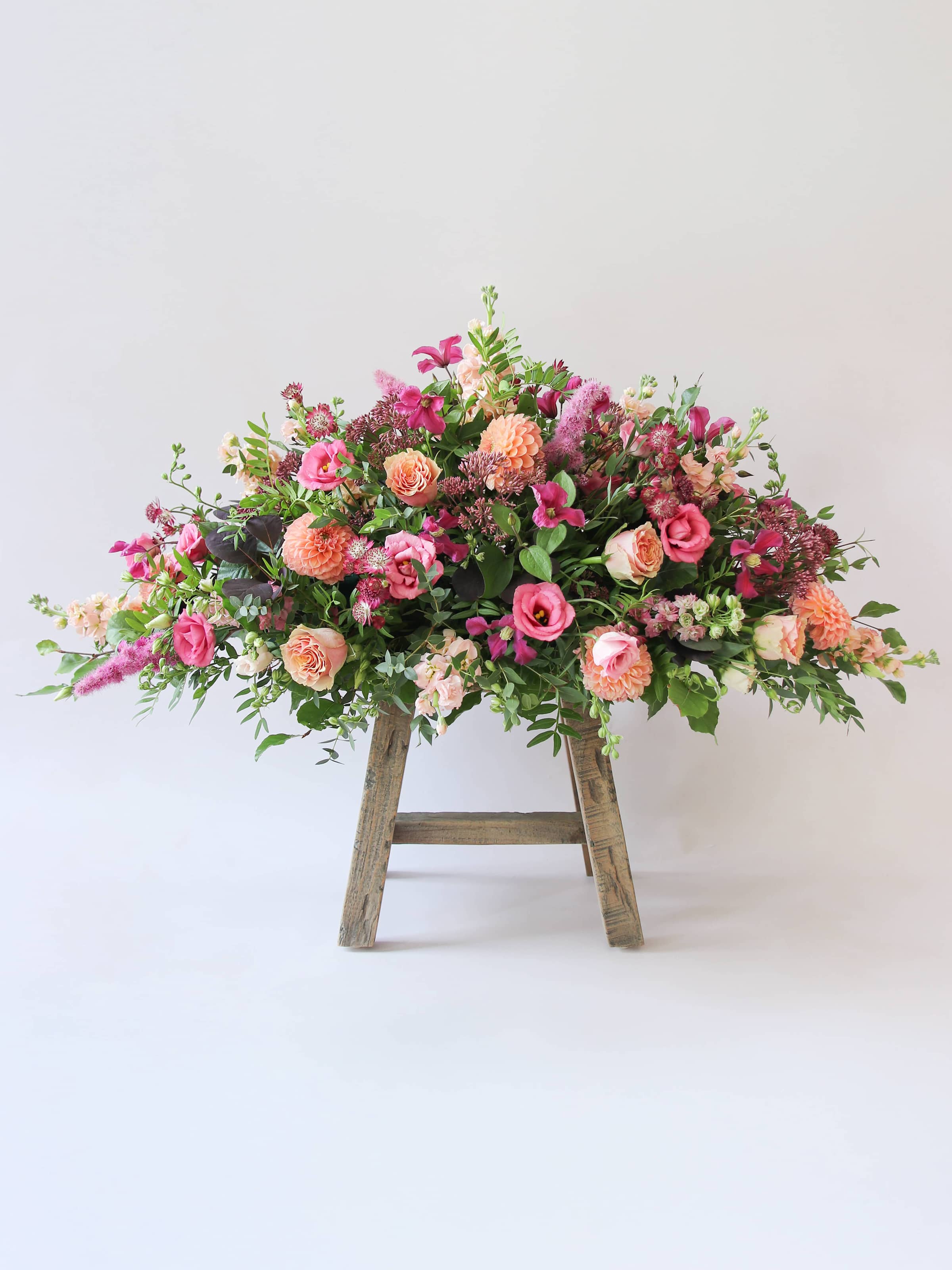 Side view of a double ended coffin spray with bright pink and peach flowers and green foliage, sitting a top a wooden stool on a white background.