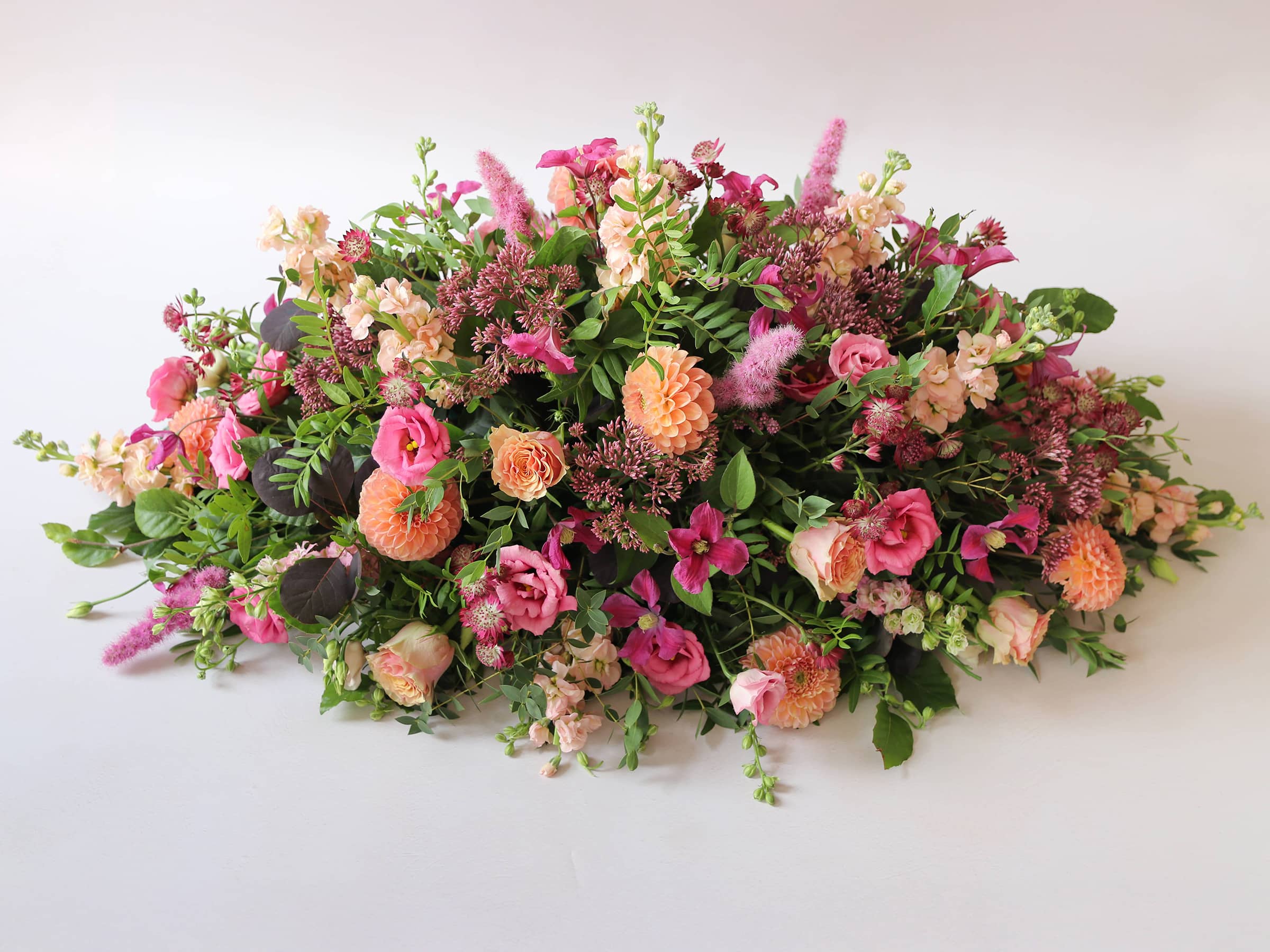 Side view of a double ended coffin spray with bright pink and peach flowers and green foliage, laying on a white background.