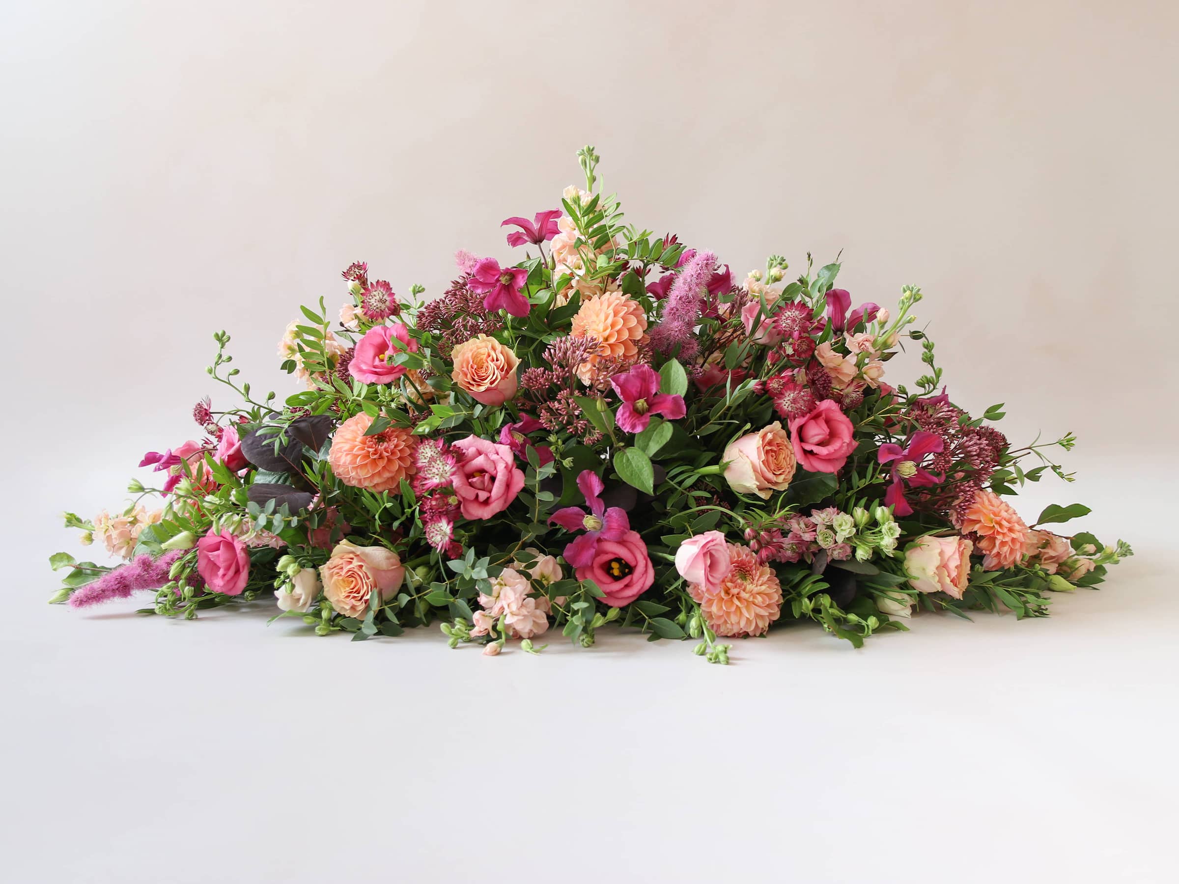 Side view of a double ended coffin spray with bright pink and peach flowers and green foliage, laying on a white background.