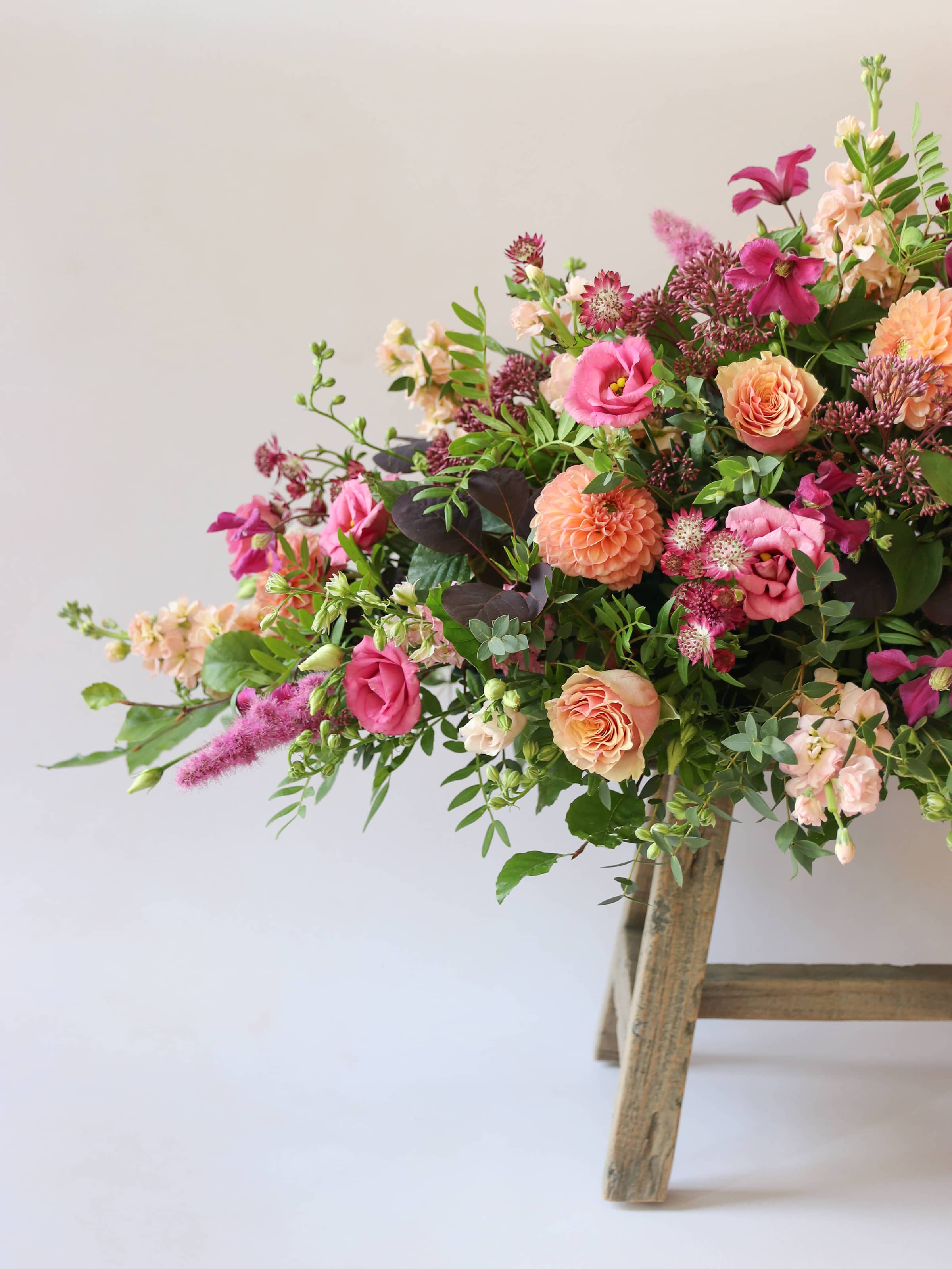 One end of a bright pink and peach coffin spray on a wooden stool against a white background