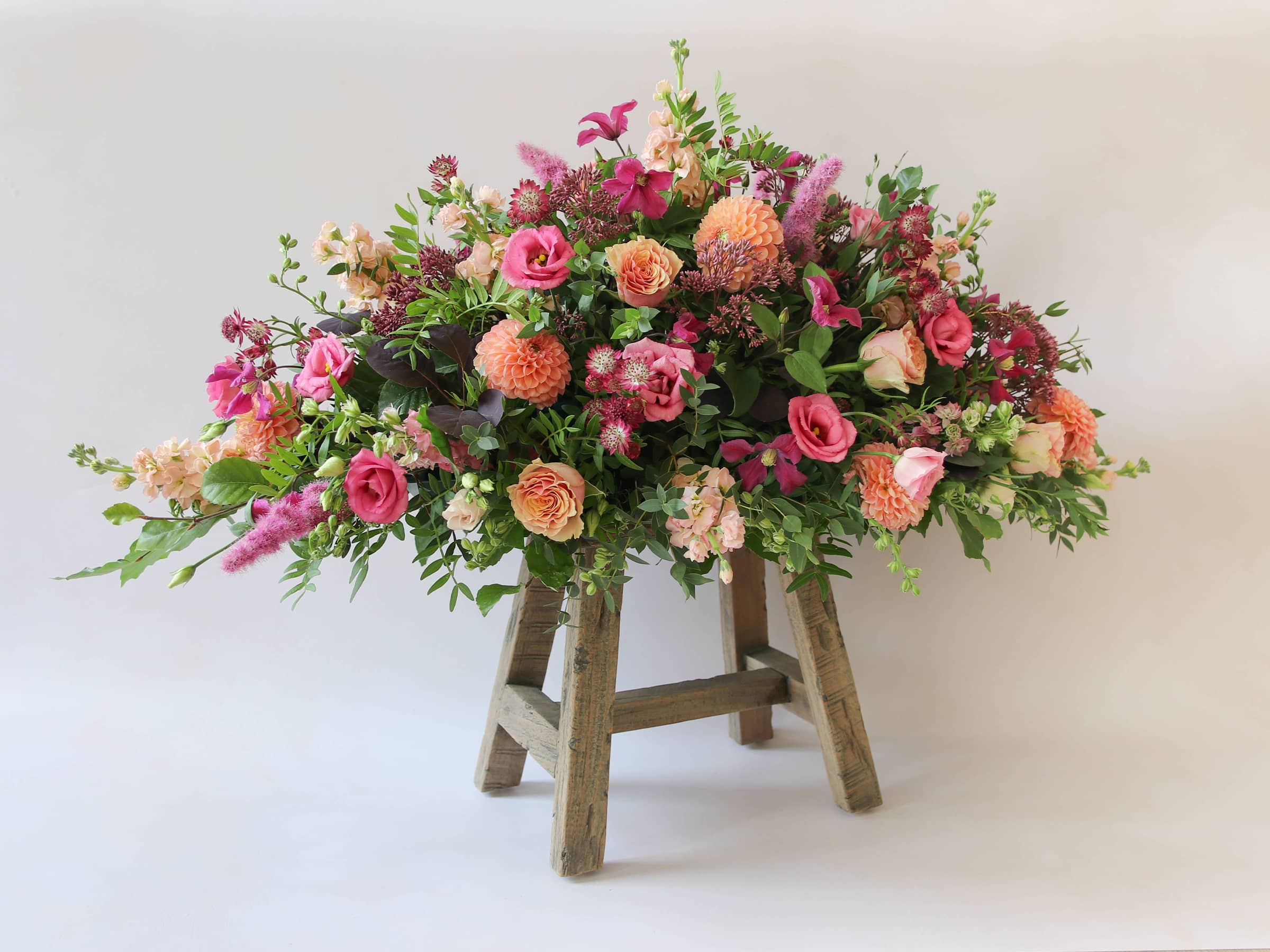 Double ended coffin spray with bright pink and peach flowers and green foliage, viewed at an angle on a wooden stool.