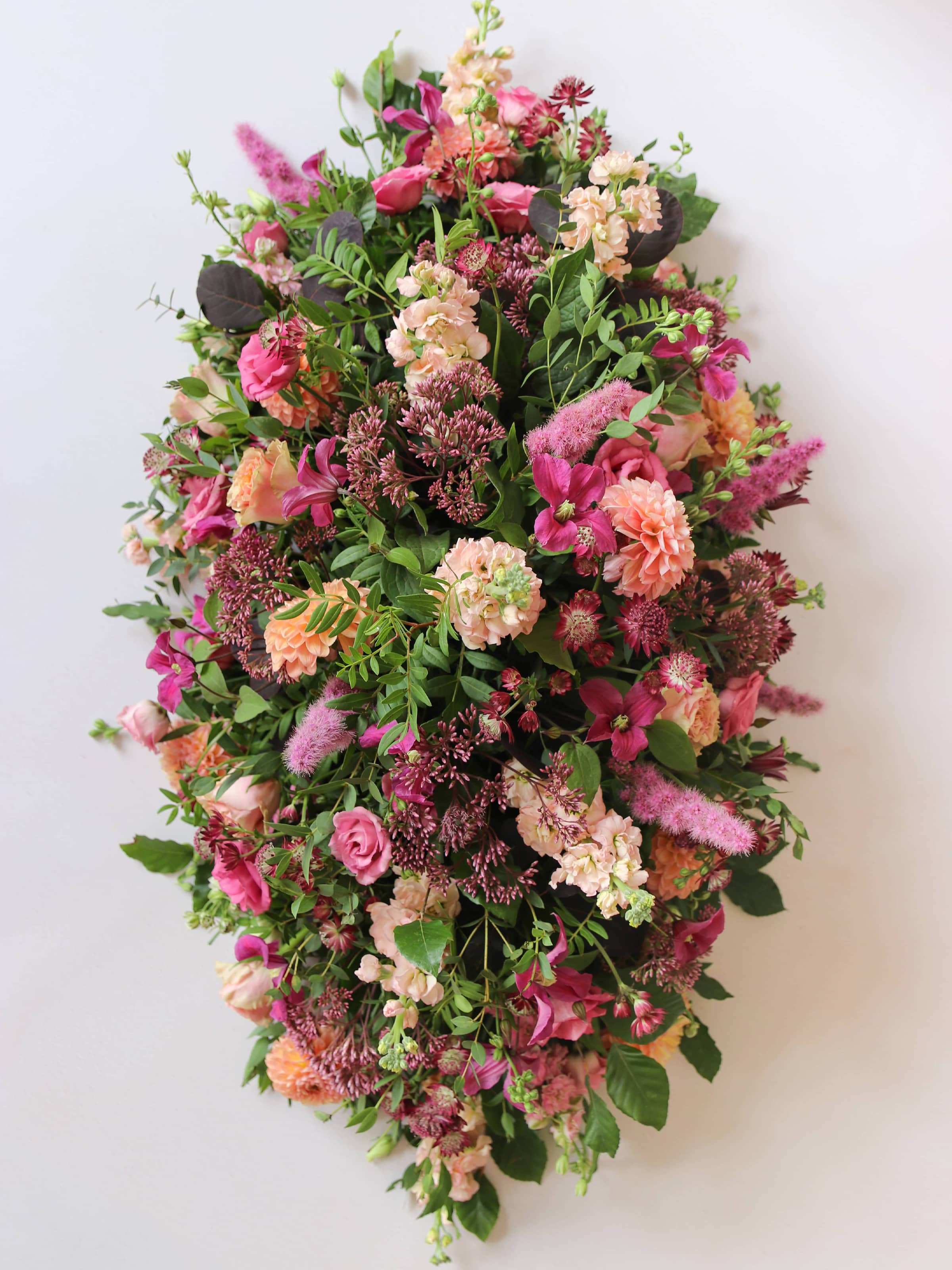 Top down view of a bright pink and peach double ended coffin spray with greenery on a white background