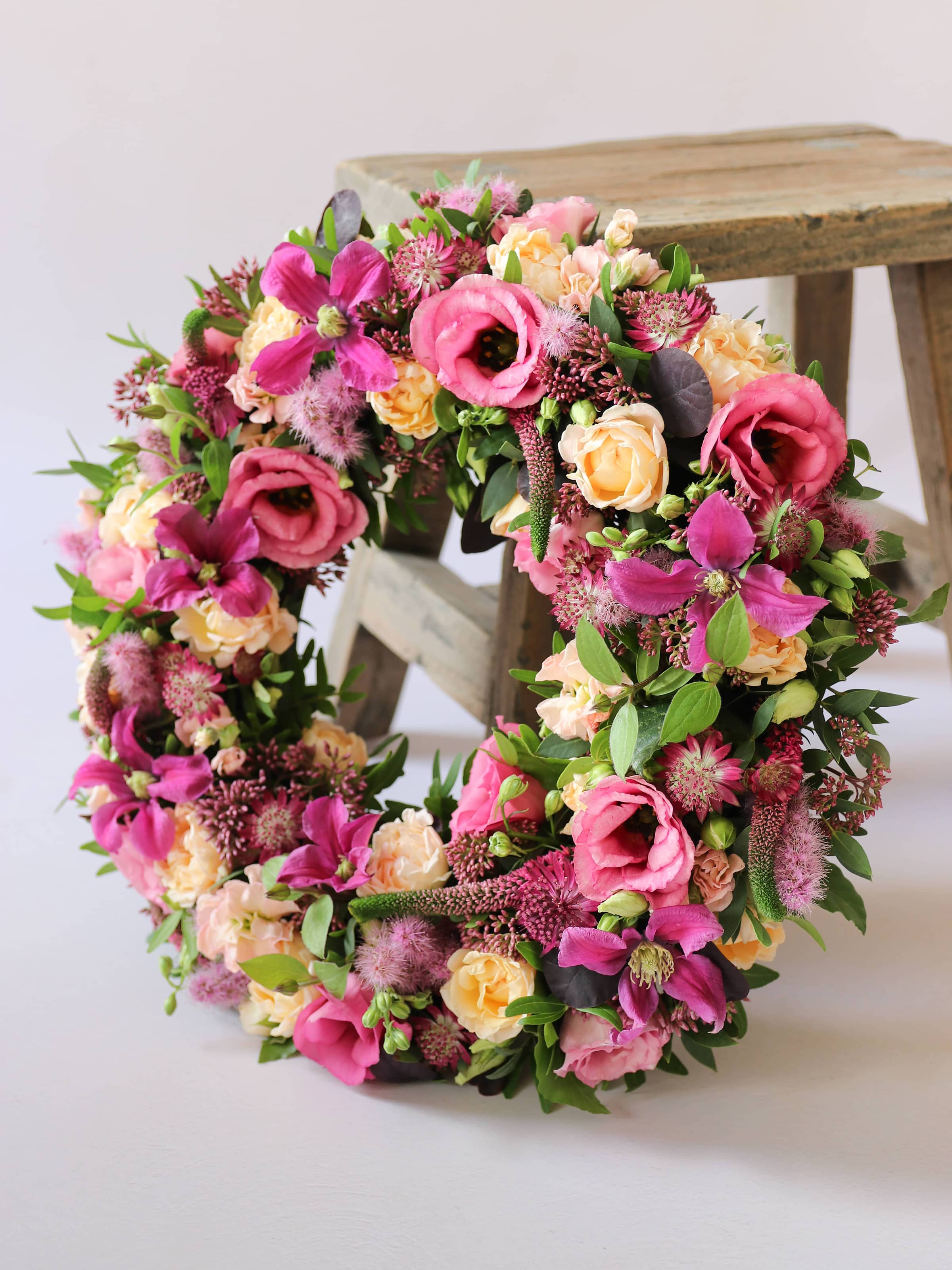 Floral wreath with pink, yellow, and green flowers on a white background with a wooden stool in the background.