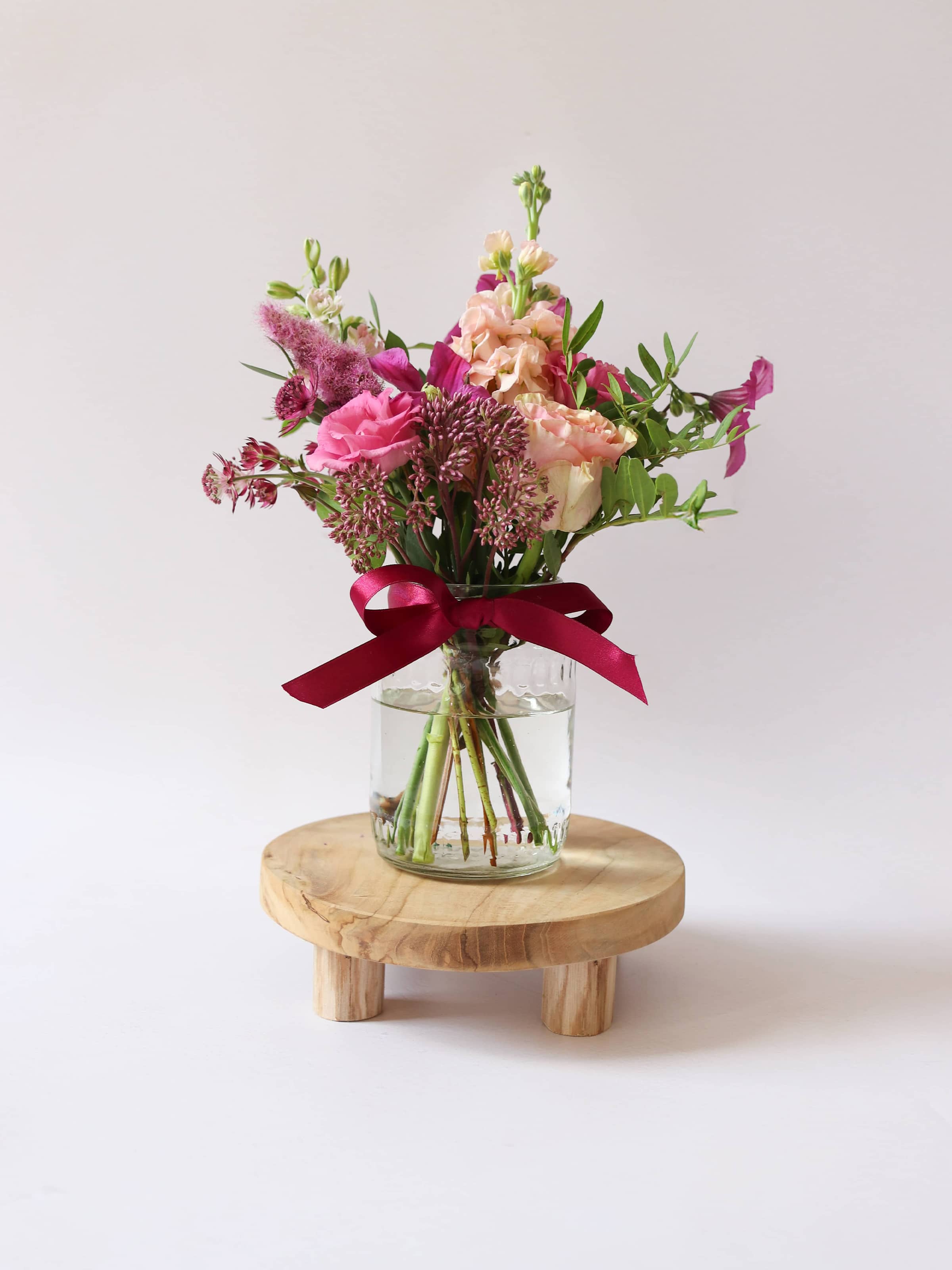 Small bouquet of flowers in a clear vase with a red ribbon on a wooden stand against a light background