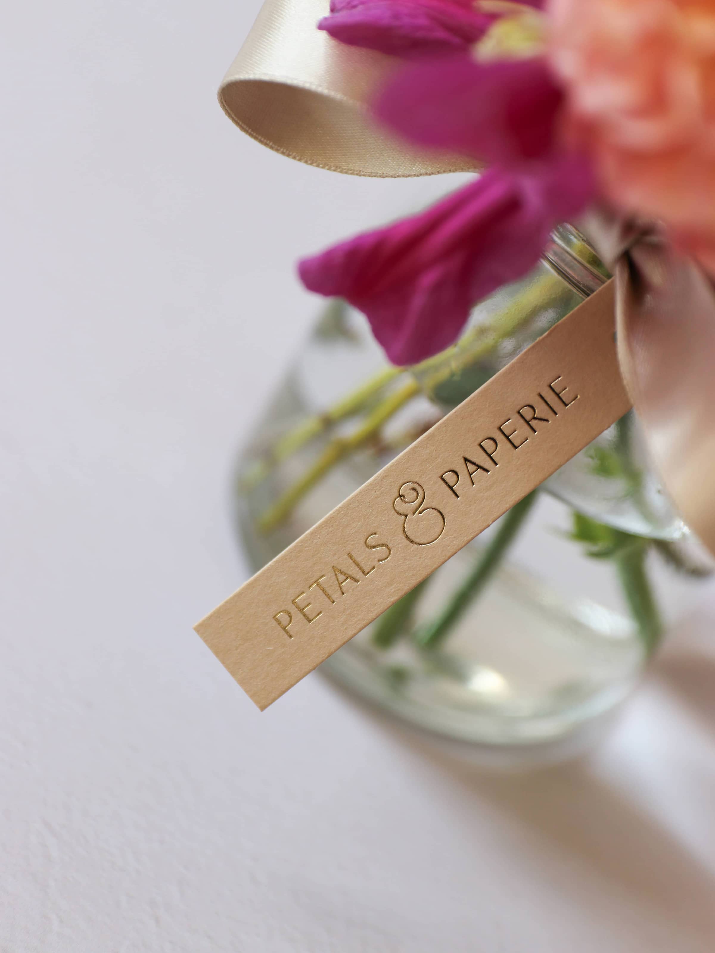 Glass vase with flowers and a 'Petals & Paperie' tag on a light background