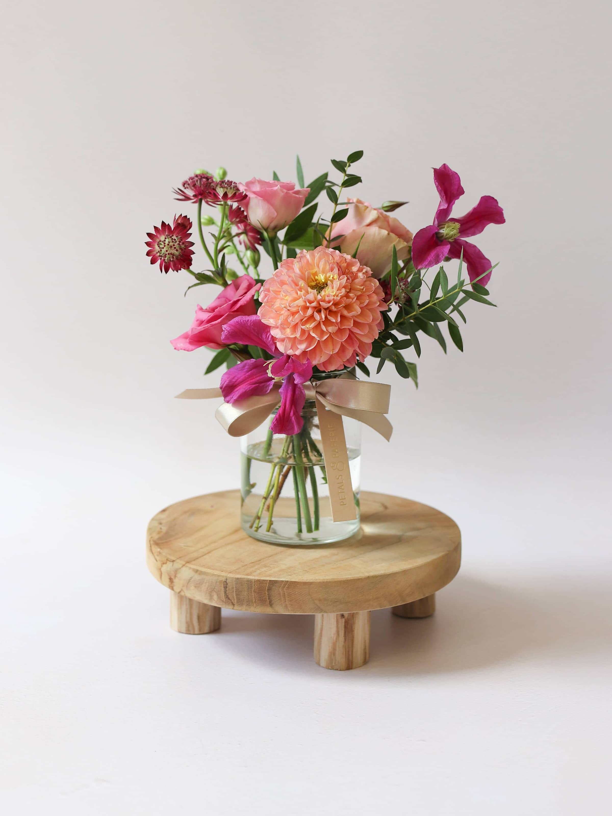 A single jam jar arrangement of bright pink and peach flowers, with a silk ribbon on top of a small wooden stool