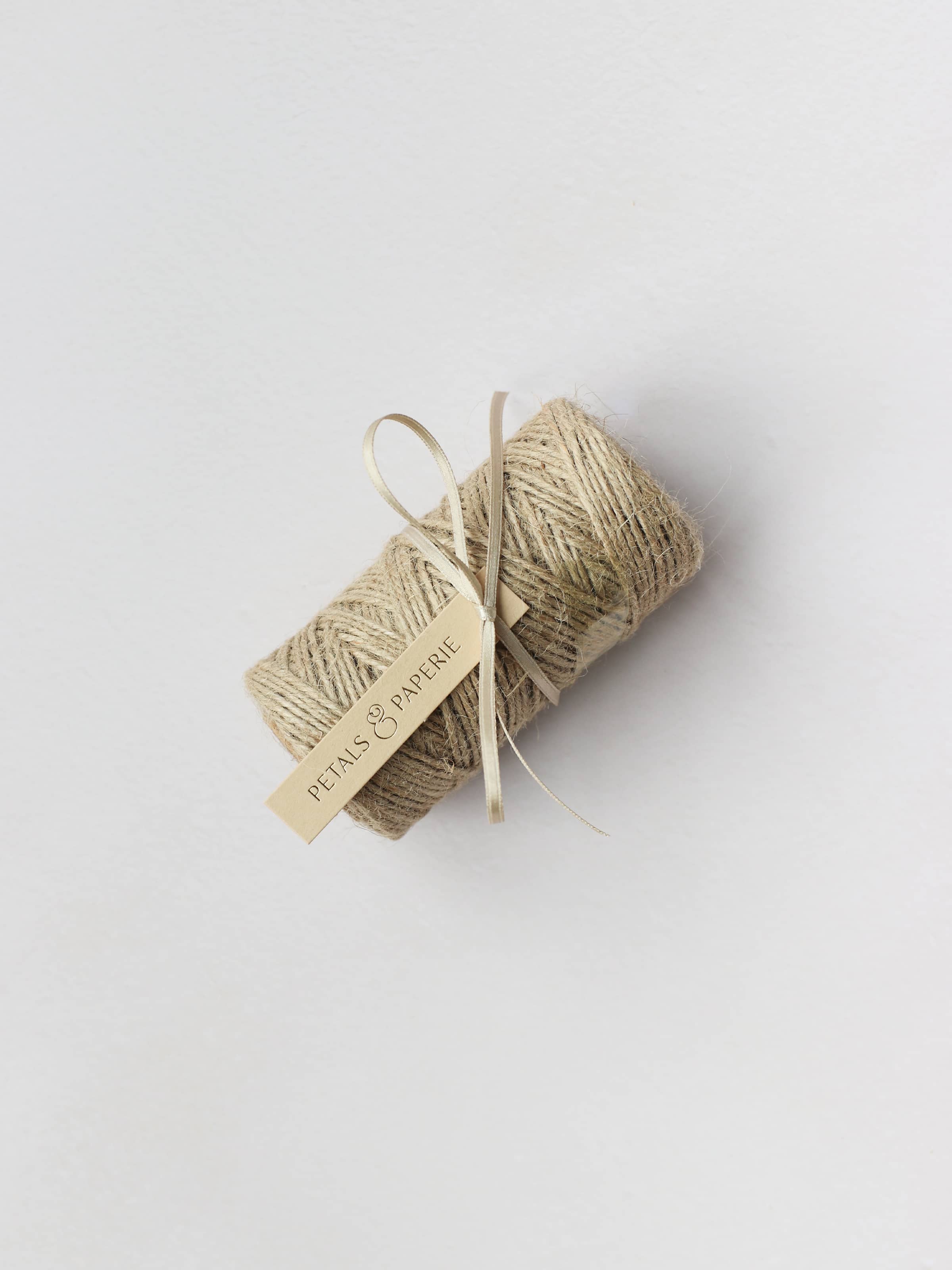 A spool of brown kraft mossing twine tied with a cream satin ribbon and a Petals & Paperie gift tag, sitting on a neutral background.