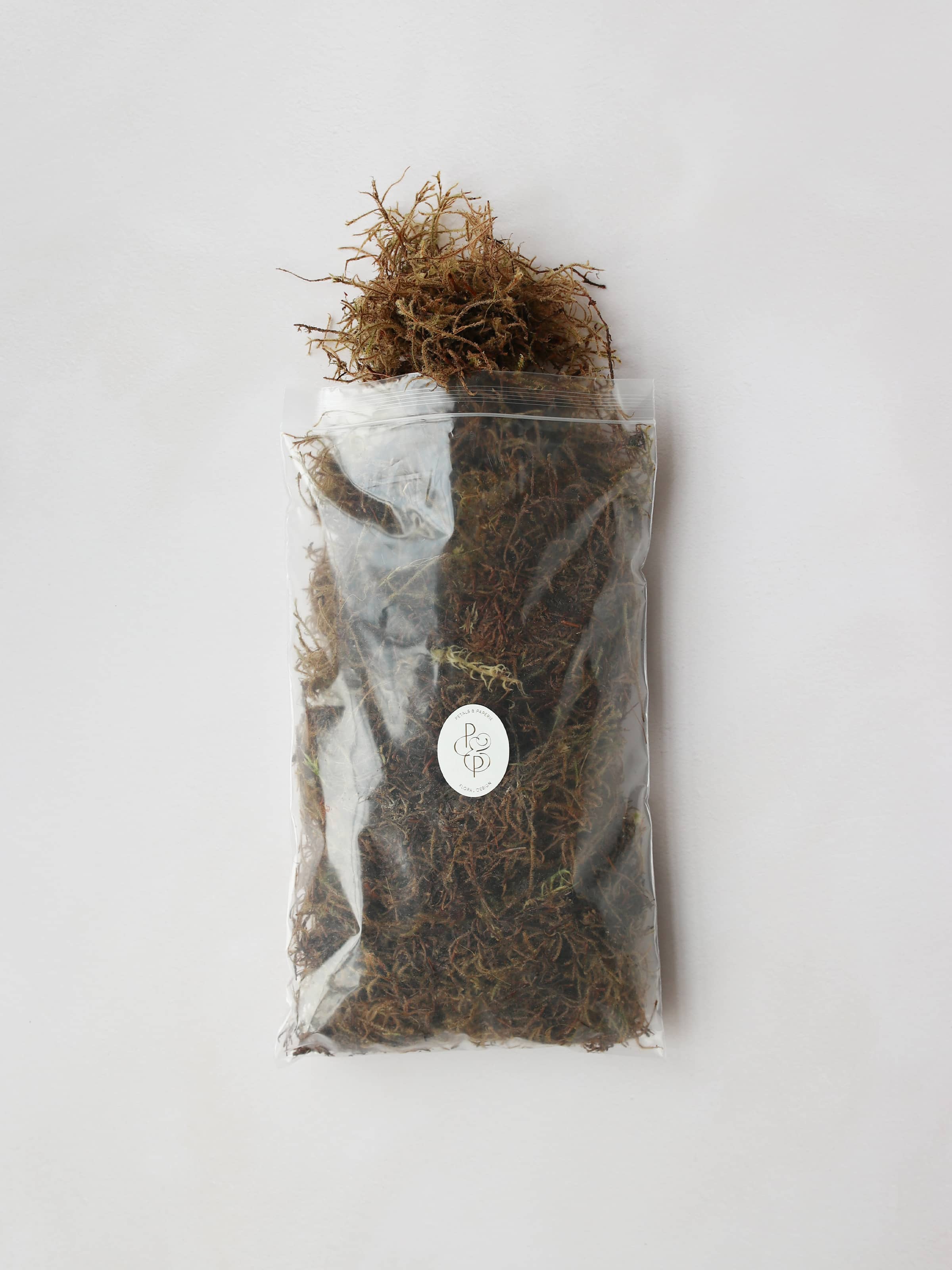 A bag of sphagnum moss packaged in an air-tight plastic resealable bag. The bag has a branded Petals & Paperie sticker on the front and the moss is trailing out of the top of the open bag.