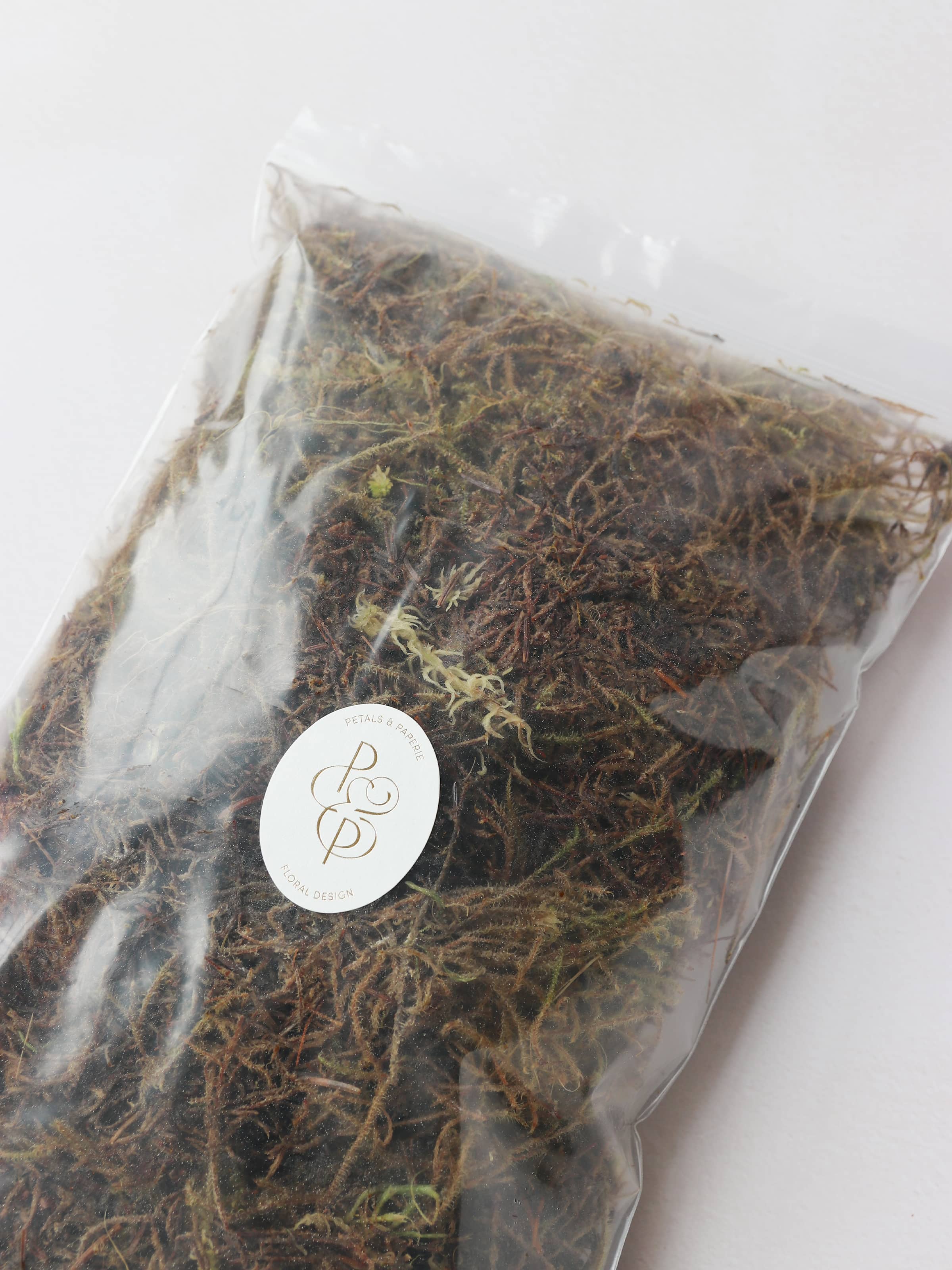 A bag of sphagnum moss packaged in an air-tight plastic resealable bag. The bag has a branded Petals & Paperie sticker on the front.