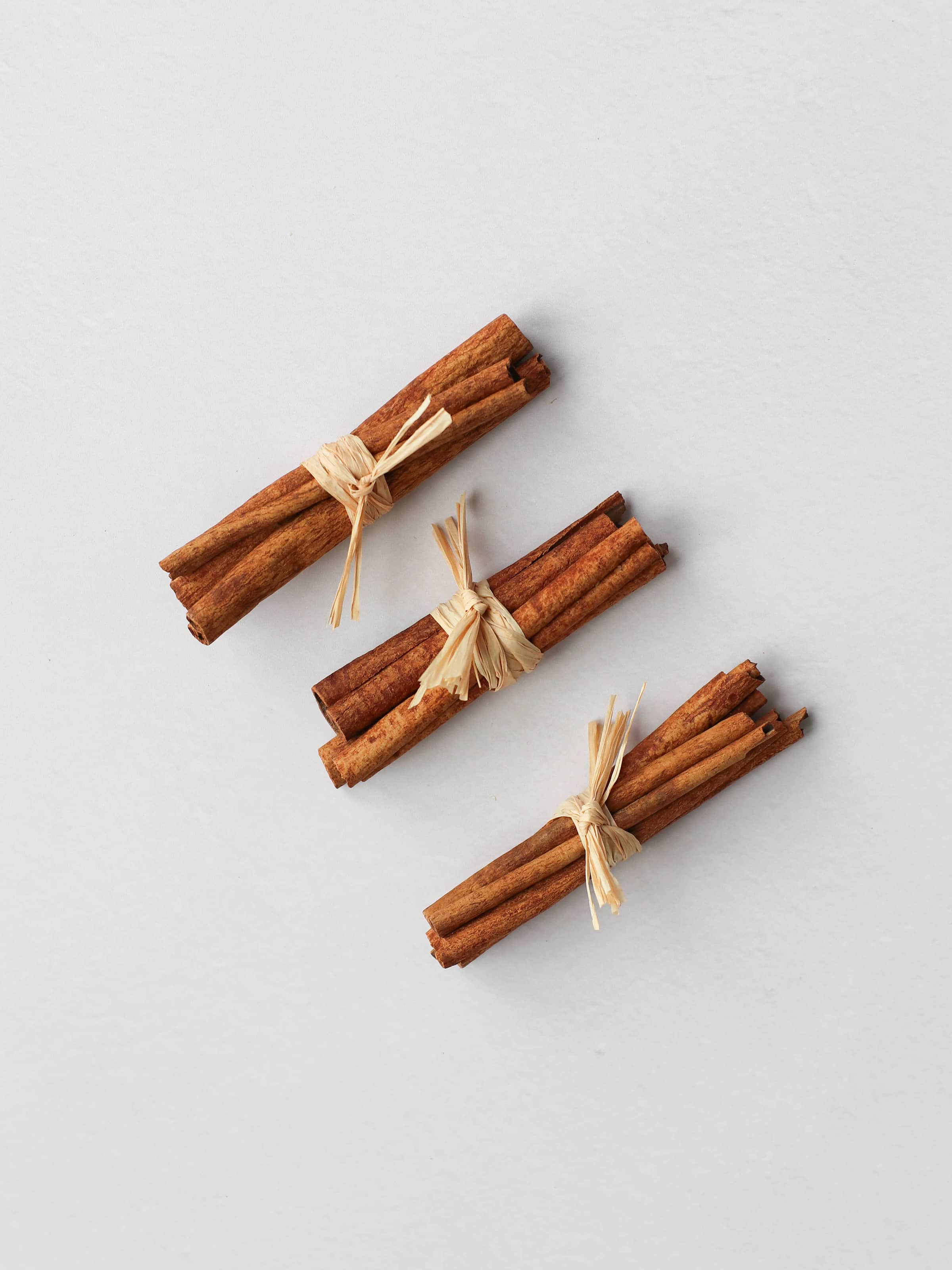 Three bundles of cinnamon sticks tied with raffia for Christmas wreath making, positioned in a diagonal row on a neutral background.