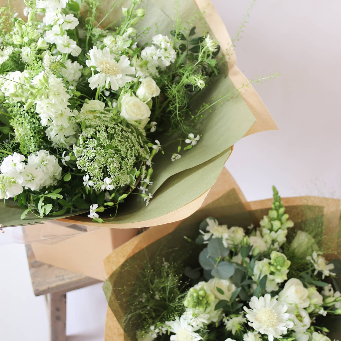 Two bouquets of white flowers with greenery gift-wrapped in brown paper wrapping on a light background.