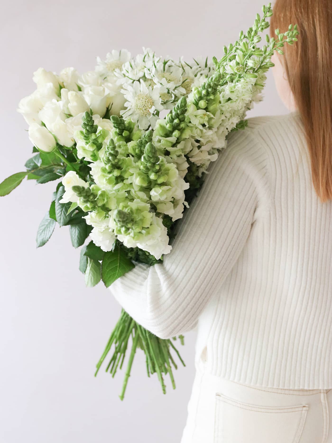 A woman wearing a white jumper and cream jeans with her back to the camera, holding a selection of white and green flowers over her shoulder against a neutral background.