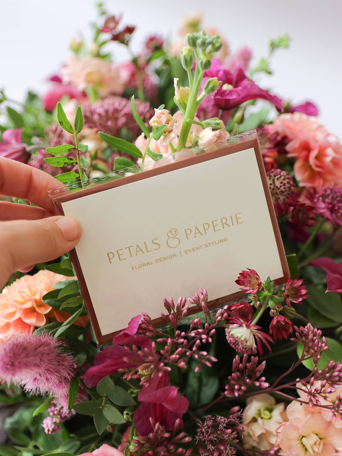 Person holding a card with 'Petals & Paperie' in a bouquet of flowers