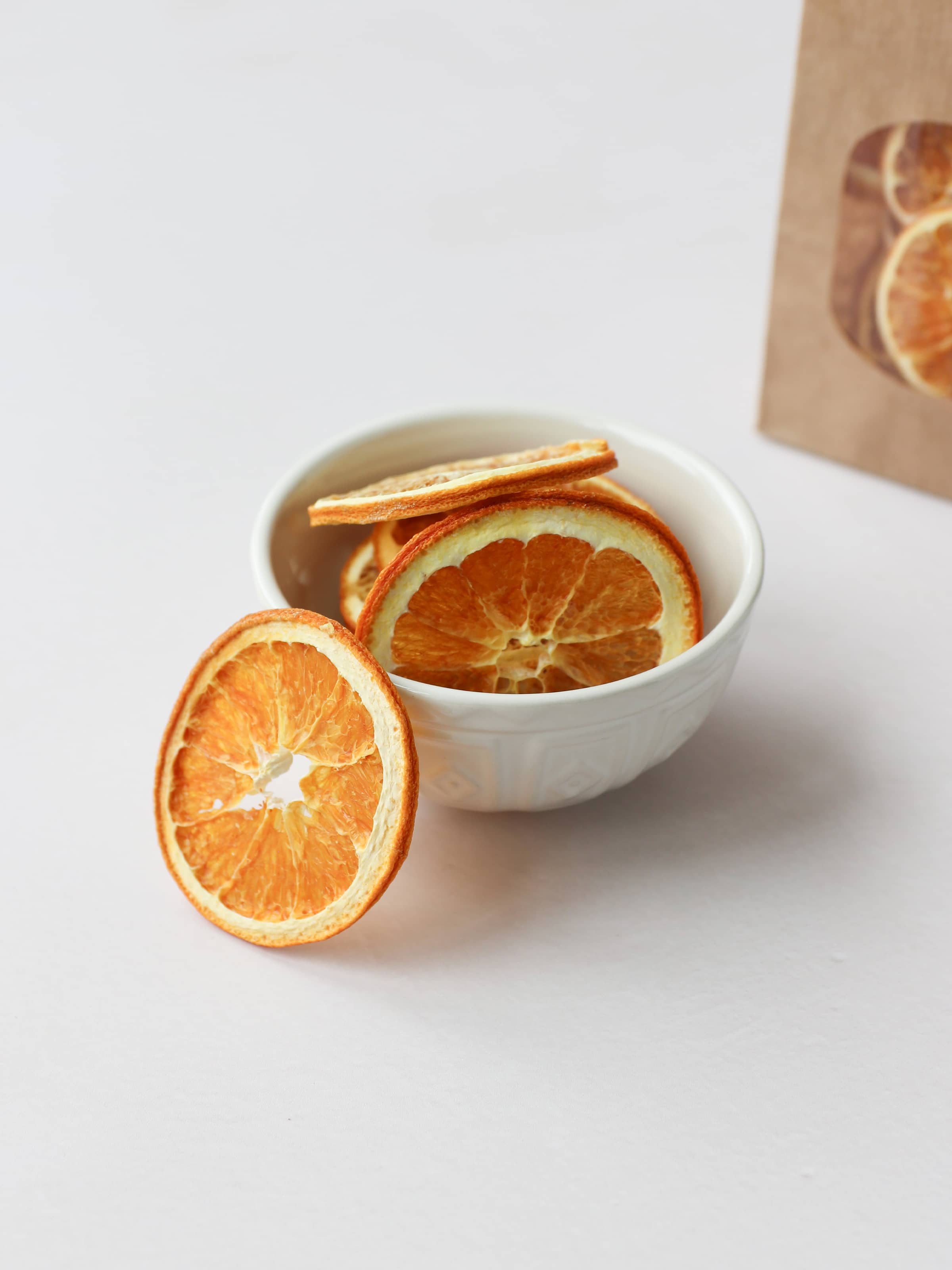 Five dried orange slices for Christmas wreath making, positioned in a cream decorative bowl, on a light background. There is a bag of dried orange slices in a brown kraft bag in the background.