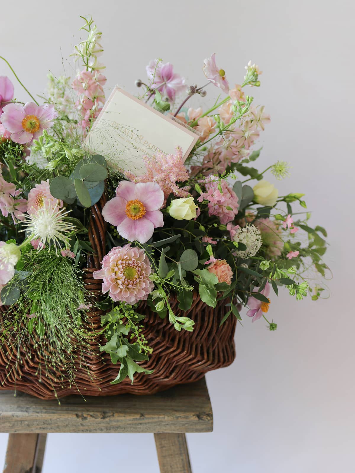 Wicker basket of pastel flowers with a gift card on a white background atop a wooden stool.