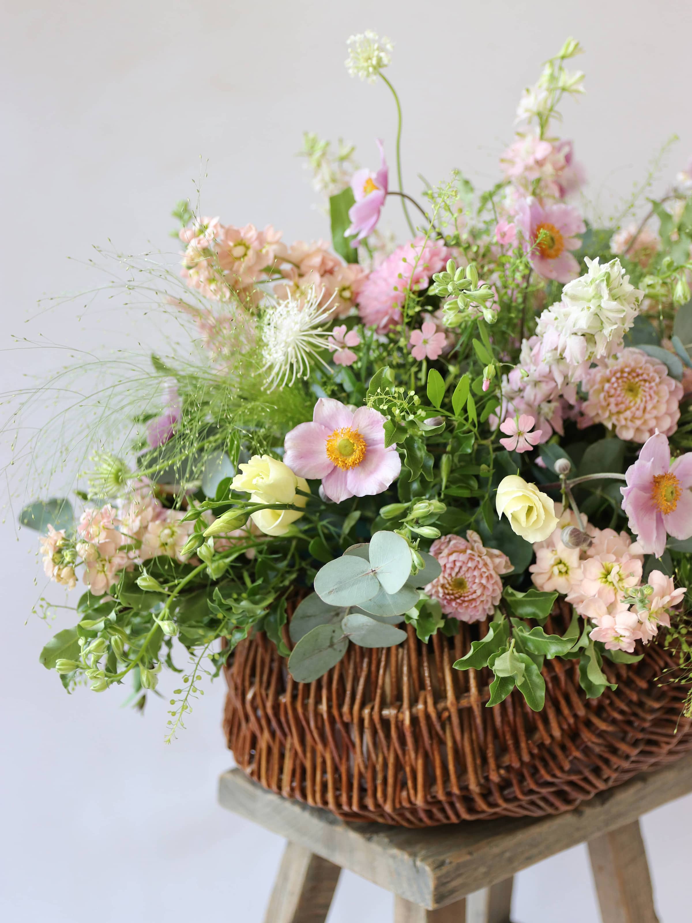 A side view of a wicker basket arrangement featuring pastel pink and yellow flowers. The basket arrangement is placed on a rustic wooden stool against a neutral background and features pink Dahlias, yellow Lisianthus and peach Stocks.
