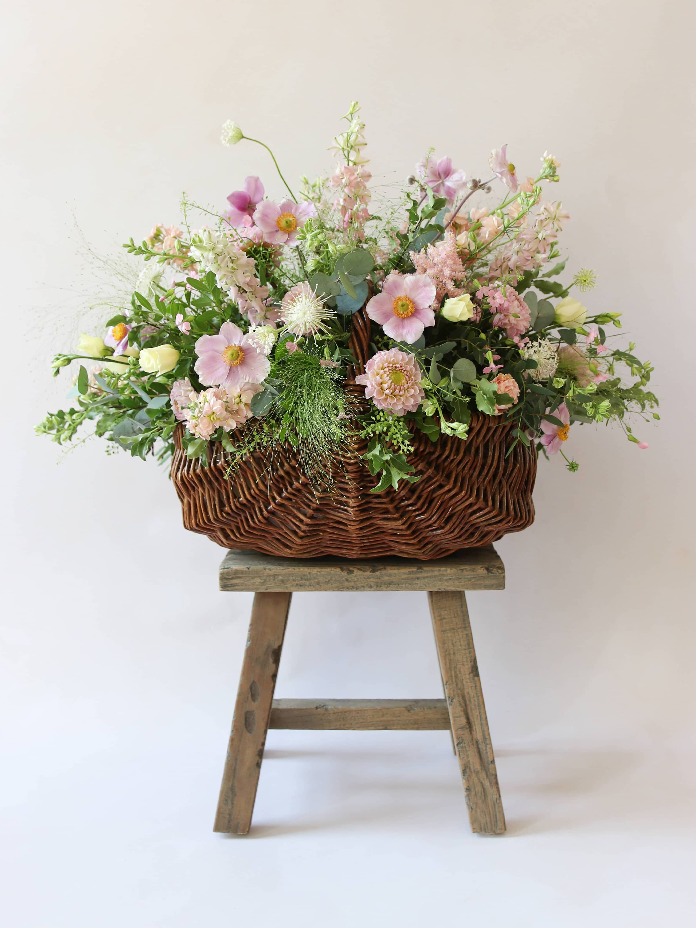 A wicker basket arrangement featuring pastel pink and yellow flowers. The basket arrangement is placed on a rustic wooden stool against a neutral background and features pink Dahlias, yellow Lisianthus and peach Stocks.