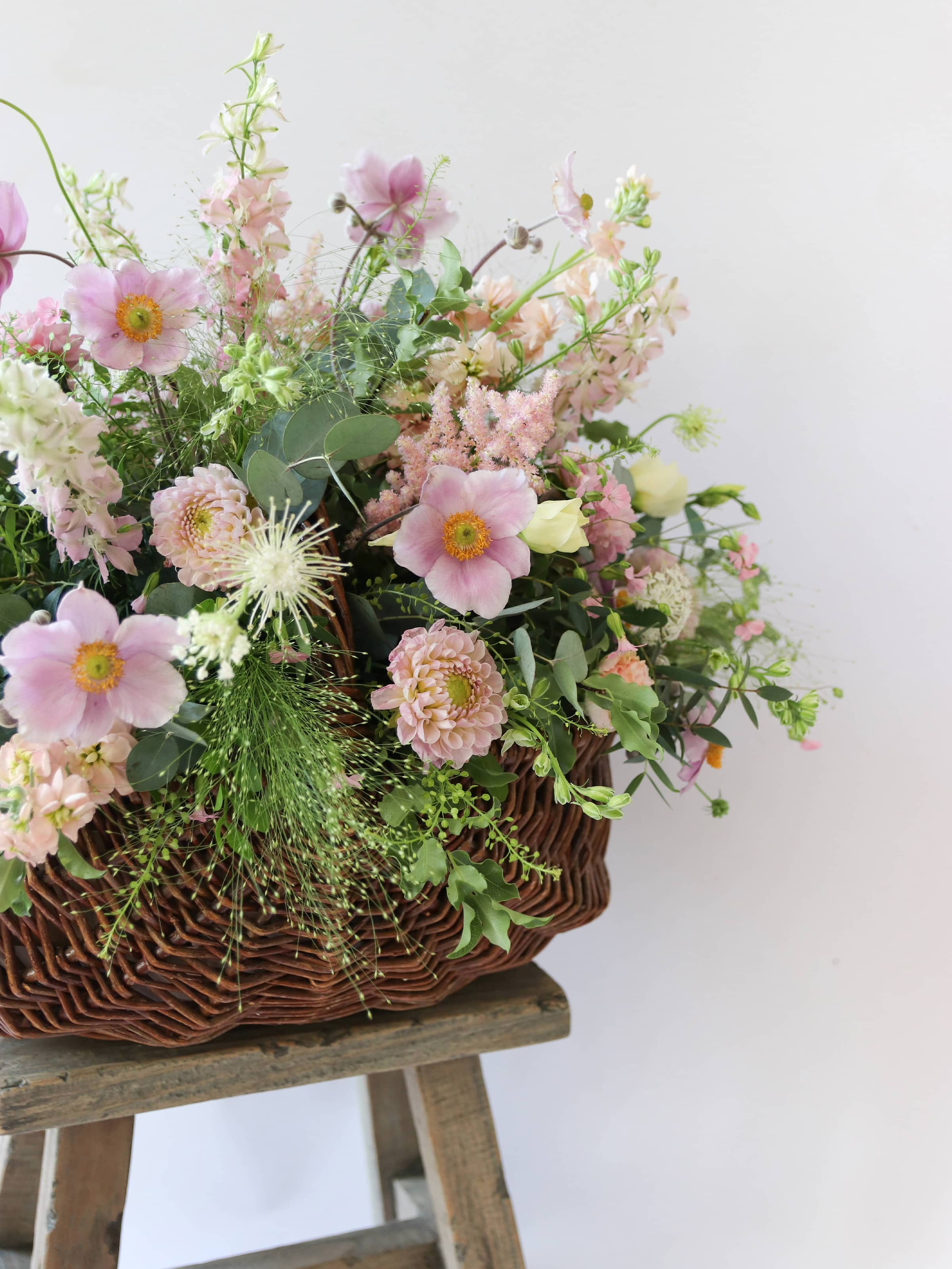 A side view of a wicker basket arrangement featuring pastel pink and yellow flowers. The basket arrangement is placed on a rustic wooden stool against a neutral background and features pink Dahlias, yellow Lisianthus and peach Stocks.