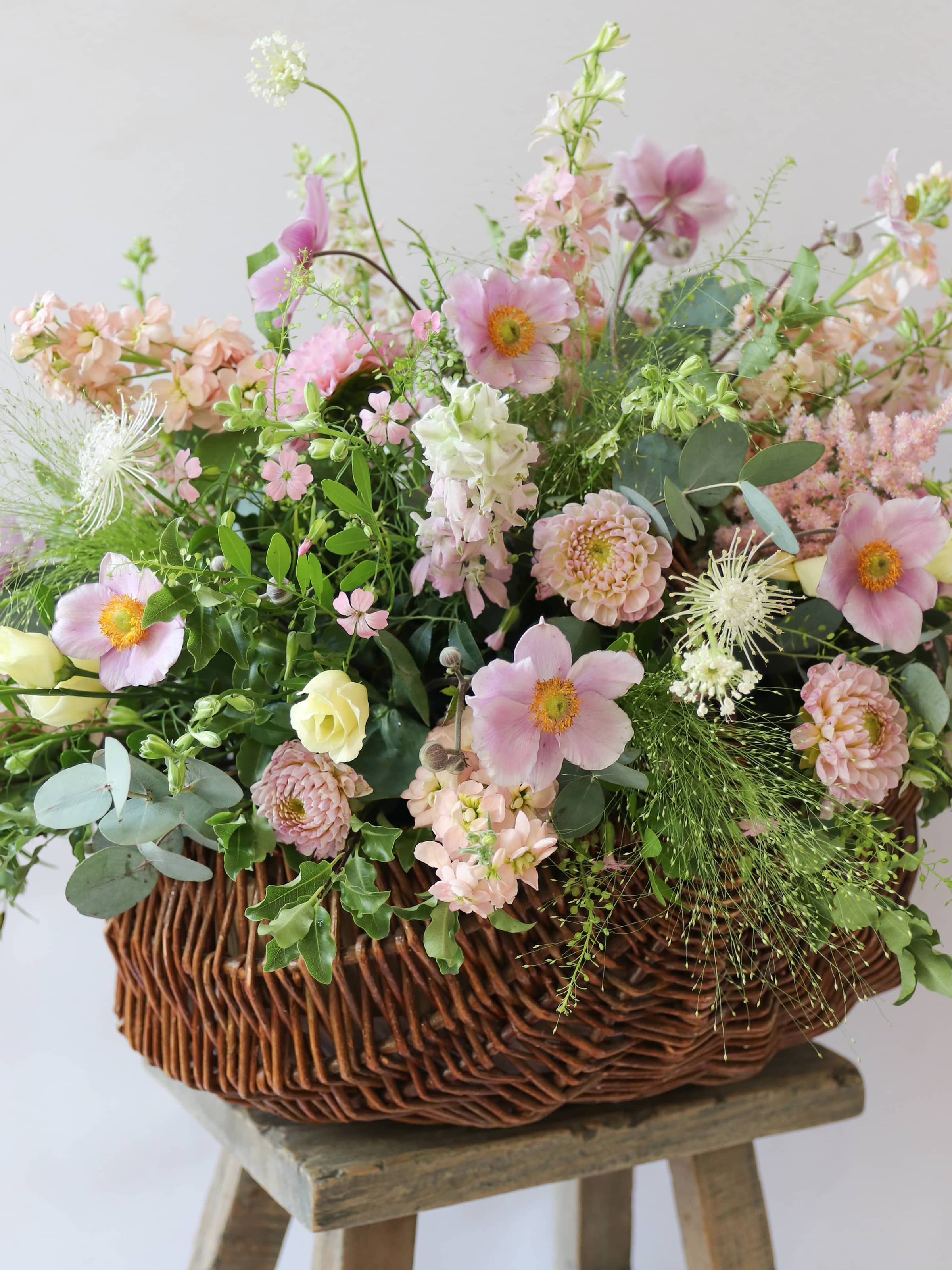 A side view of a wicker basket arrangement featuring pastel pink and yellow flowers. The basket arrangement is placed on a rustic wooden stool against a neutral background and features pink Dahlias, yellow Lisianthus and peach Stocks.