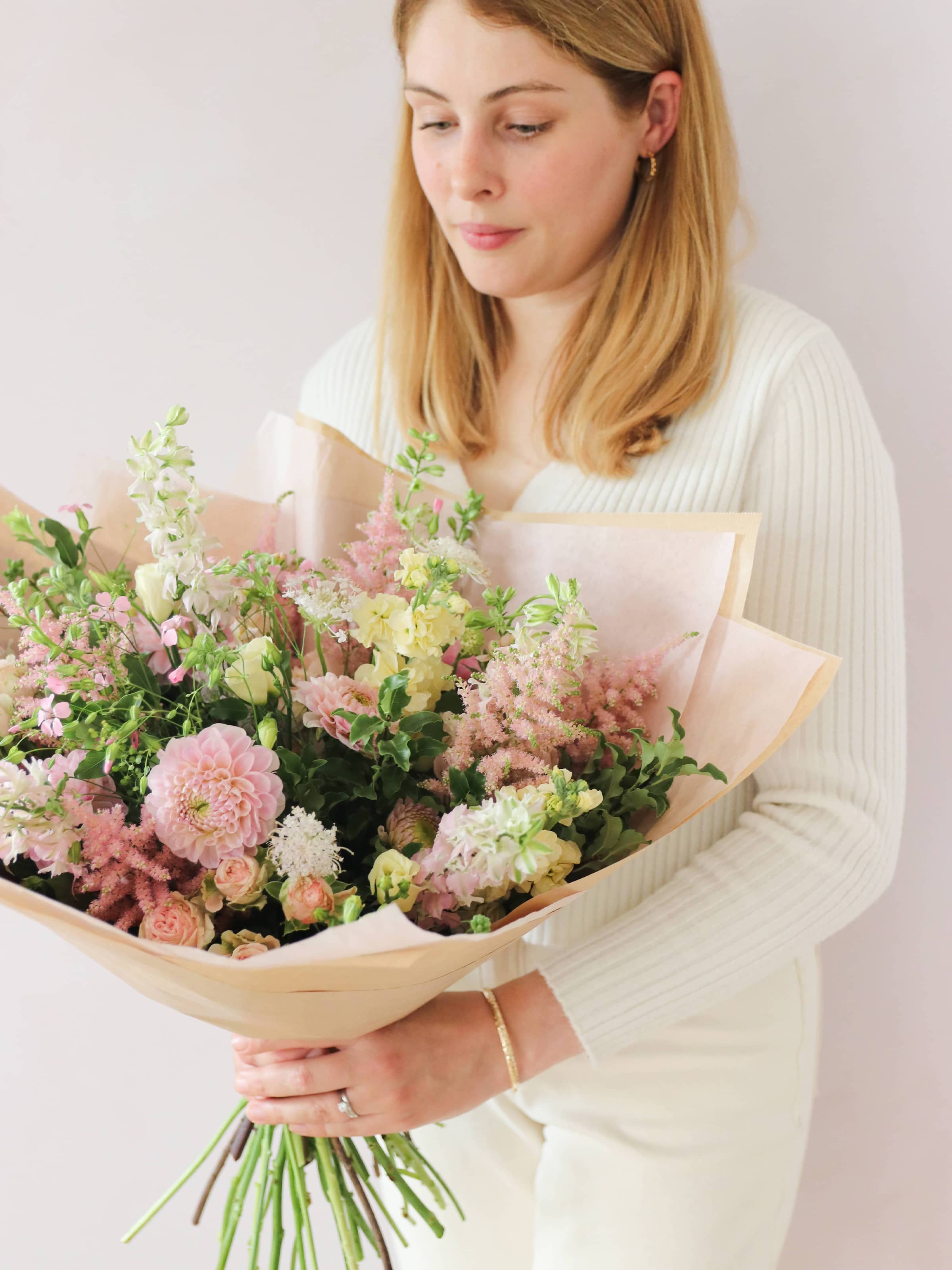 A woman standing in front of a neutral background holding a pastel yellow and pink gift-wrapped bouquet of flowers including Astilbe, Dahlias, Roses and Lisianthus in their hands.