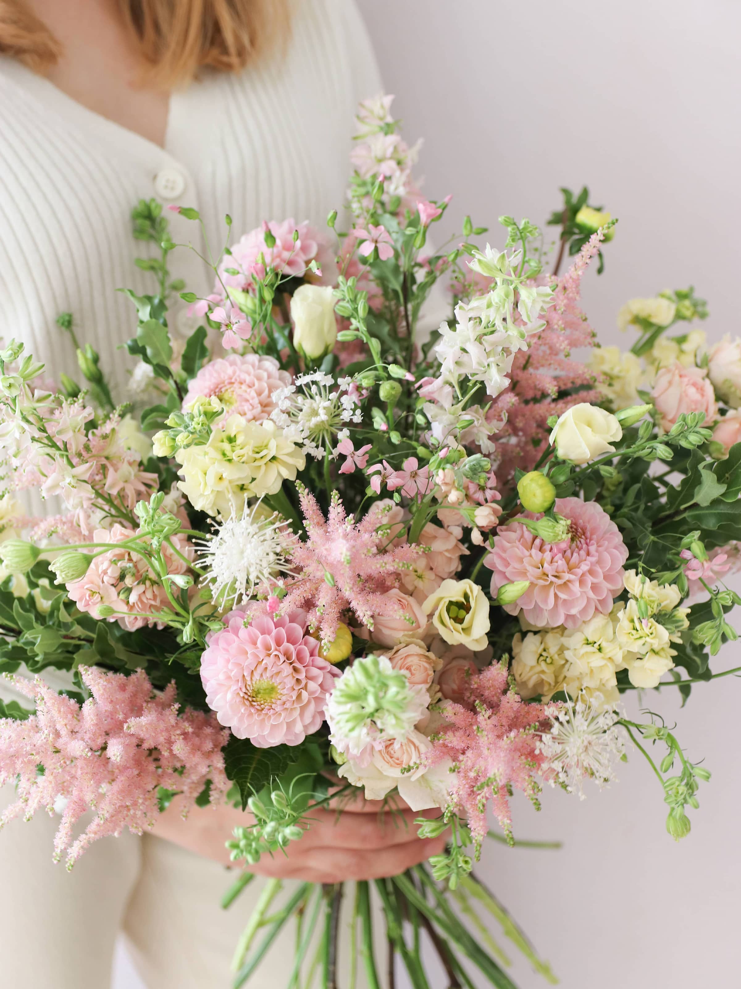 A woman wearing a white jumper and cream jeans, holding a pastel yellow and pink bouquet of flowers including Astilbe, Dahlias, Roses and Lisianthus in both hands against a neutral background.