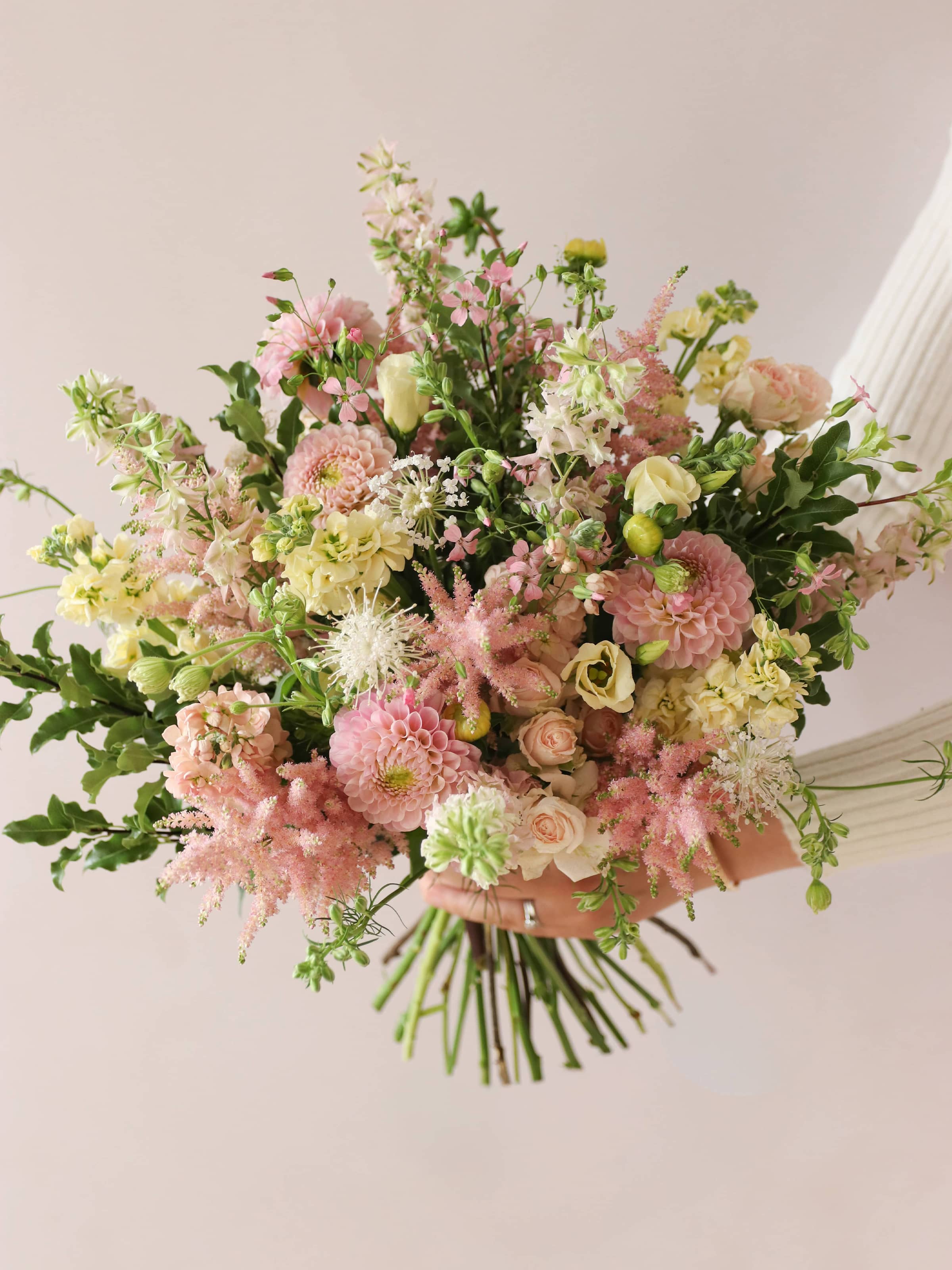 A person holding a pastel yellow and pink bouquet of flowers including Astilbe, Dahlias, Roses and Lisianthus in both hands against a neutral background.