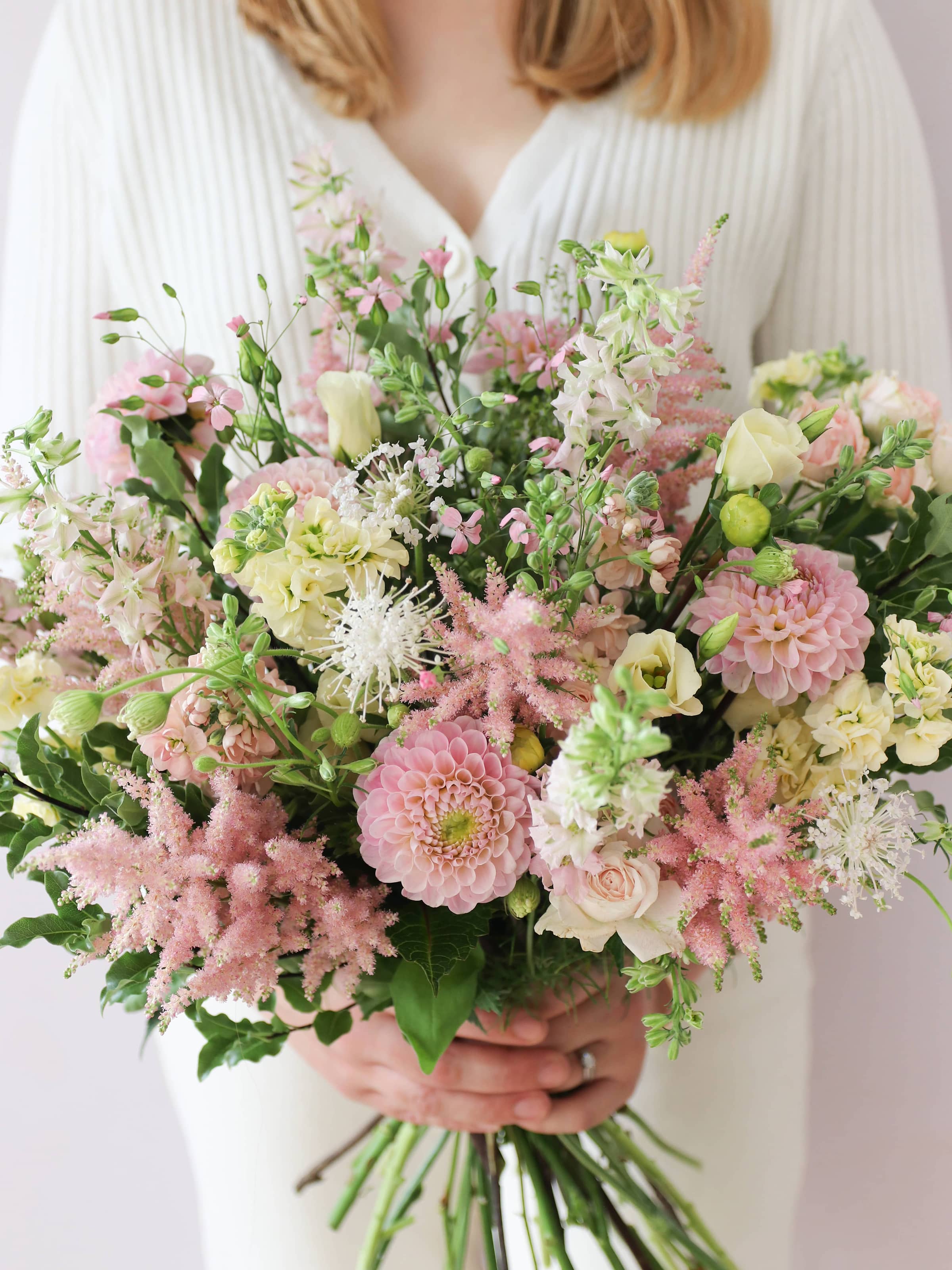 A woman wearing a white jumper and cream jeans, holding a pastel yellow and pink bouquet of flowers including Astilbe, Dahlias, Roses and Lisianthus in both hands.