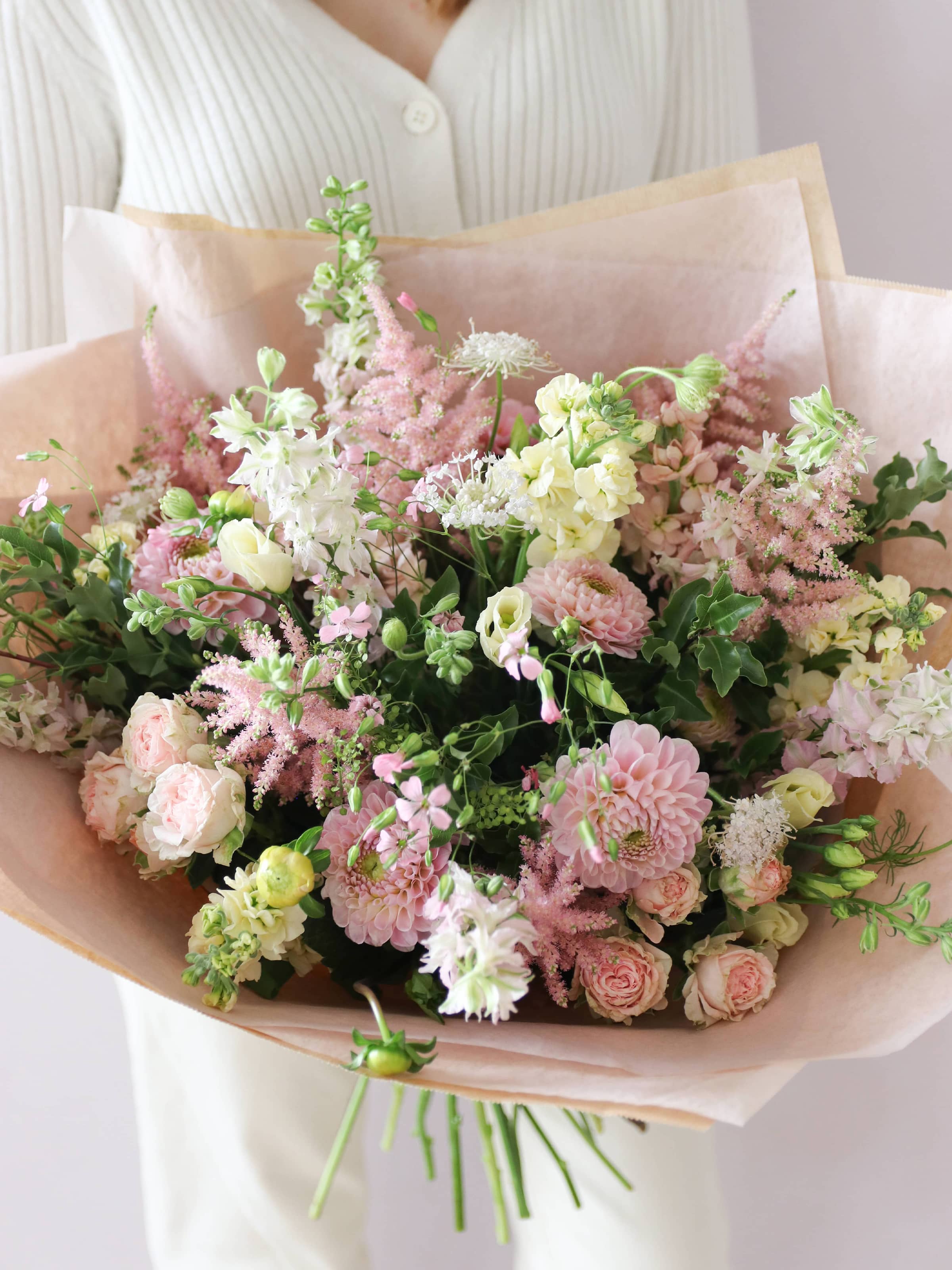 A pastel yellow and pink bouquet of flowers, gift-wrapped in brown paper on a light background, being held by a person wearing a white jumper and cream jeans.