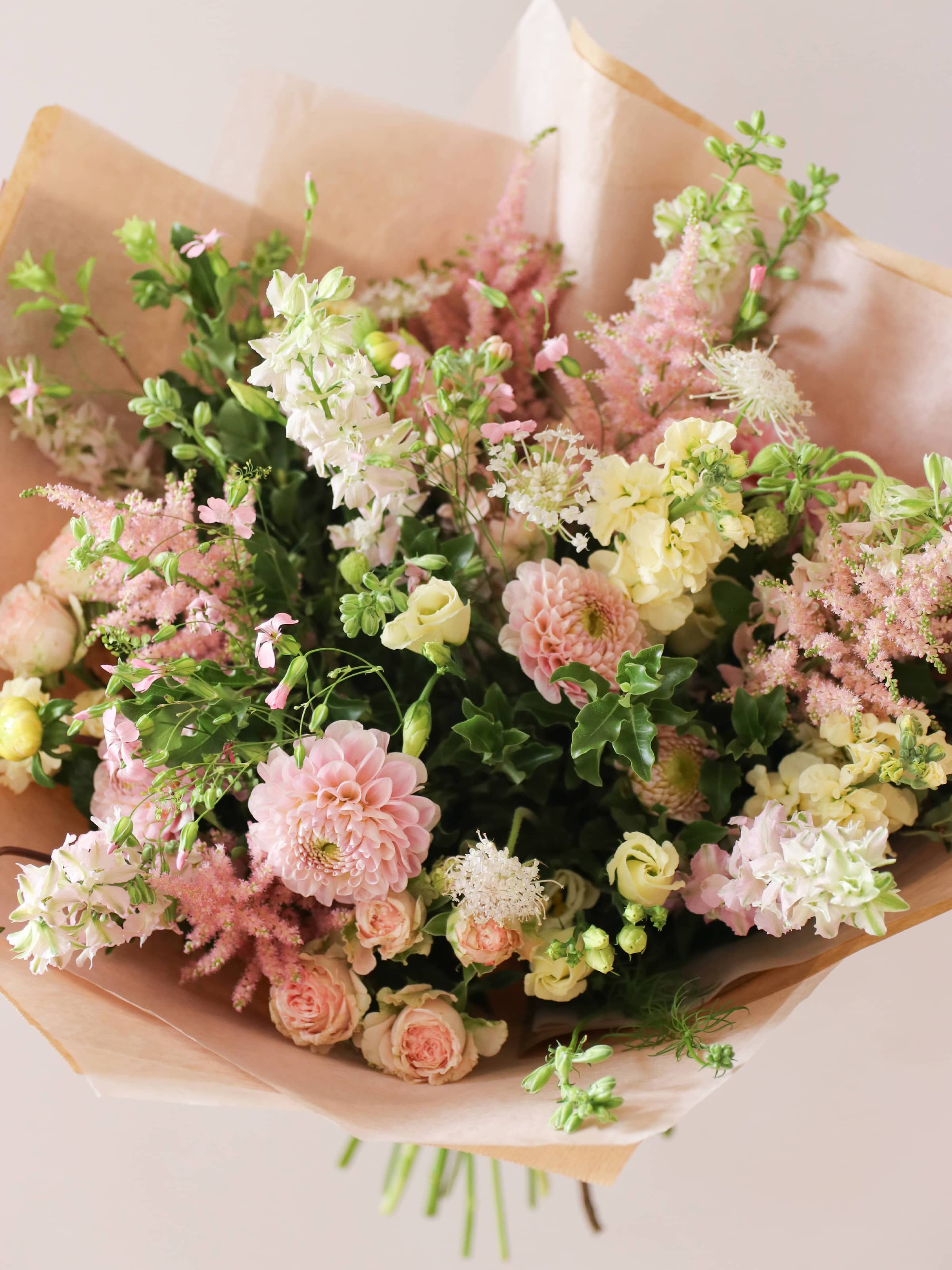 A pastel yellow and pink bouquet of flowers, gift-wrapped in brown paper on a light background.