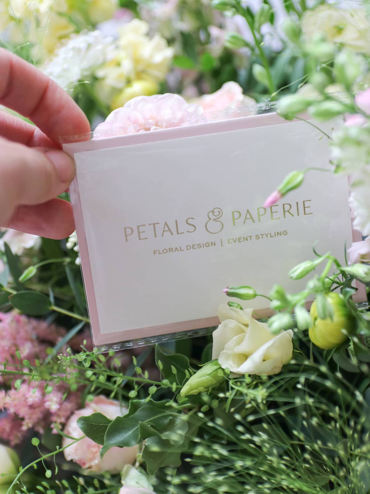 Hand holding a message card with 'Petals & Papieré' branding amidst a pastel flower arrangement.