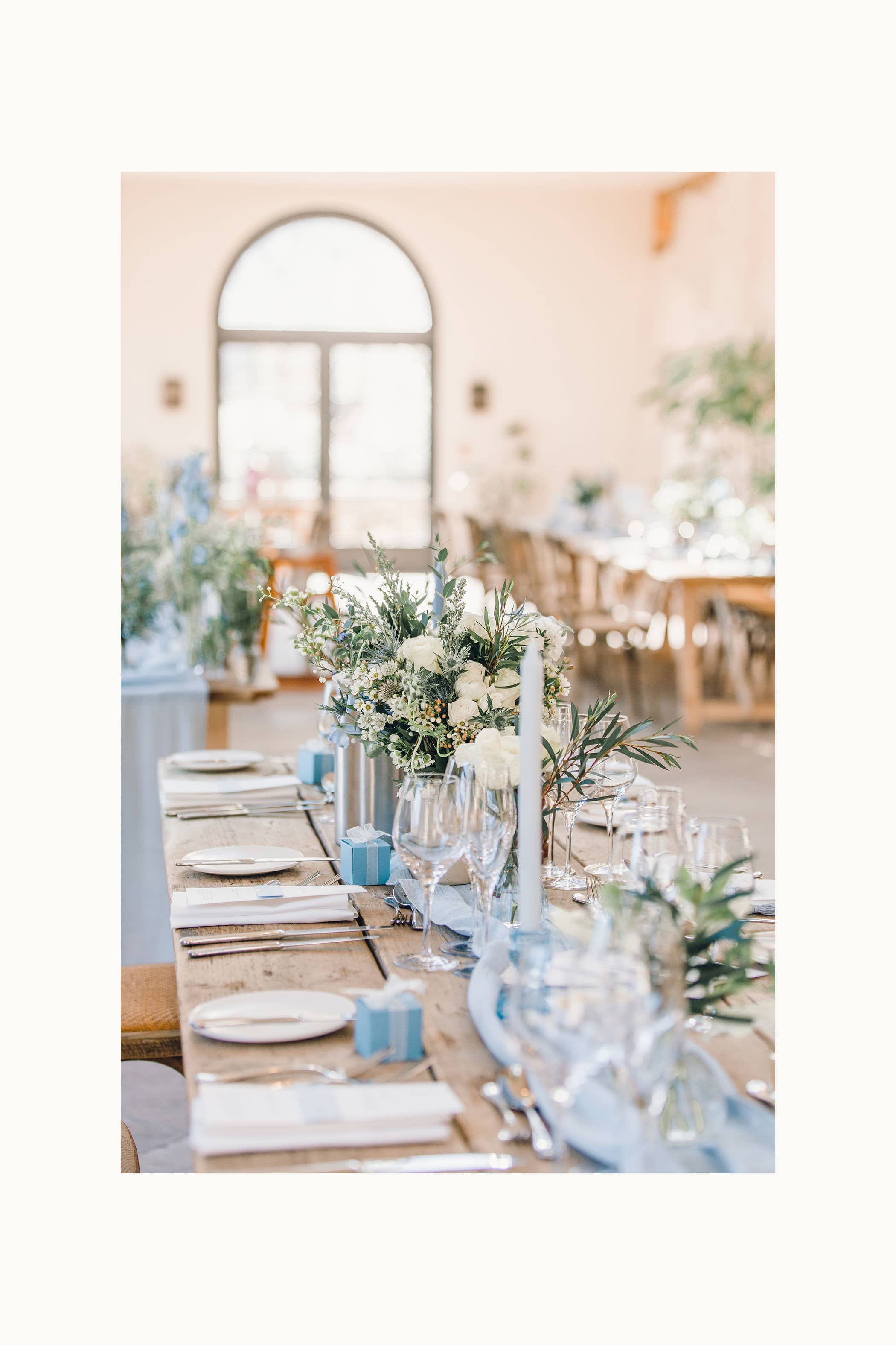 Decorative wedding table setting with white and blue floral arrangements and blue dinner candles in a softly lit room at The Fig House, Middleton Lodge. The tables include blue favour boxes, silver cutlery and bud vases full of white and blue flowers.