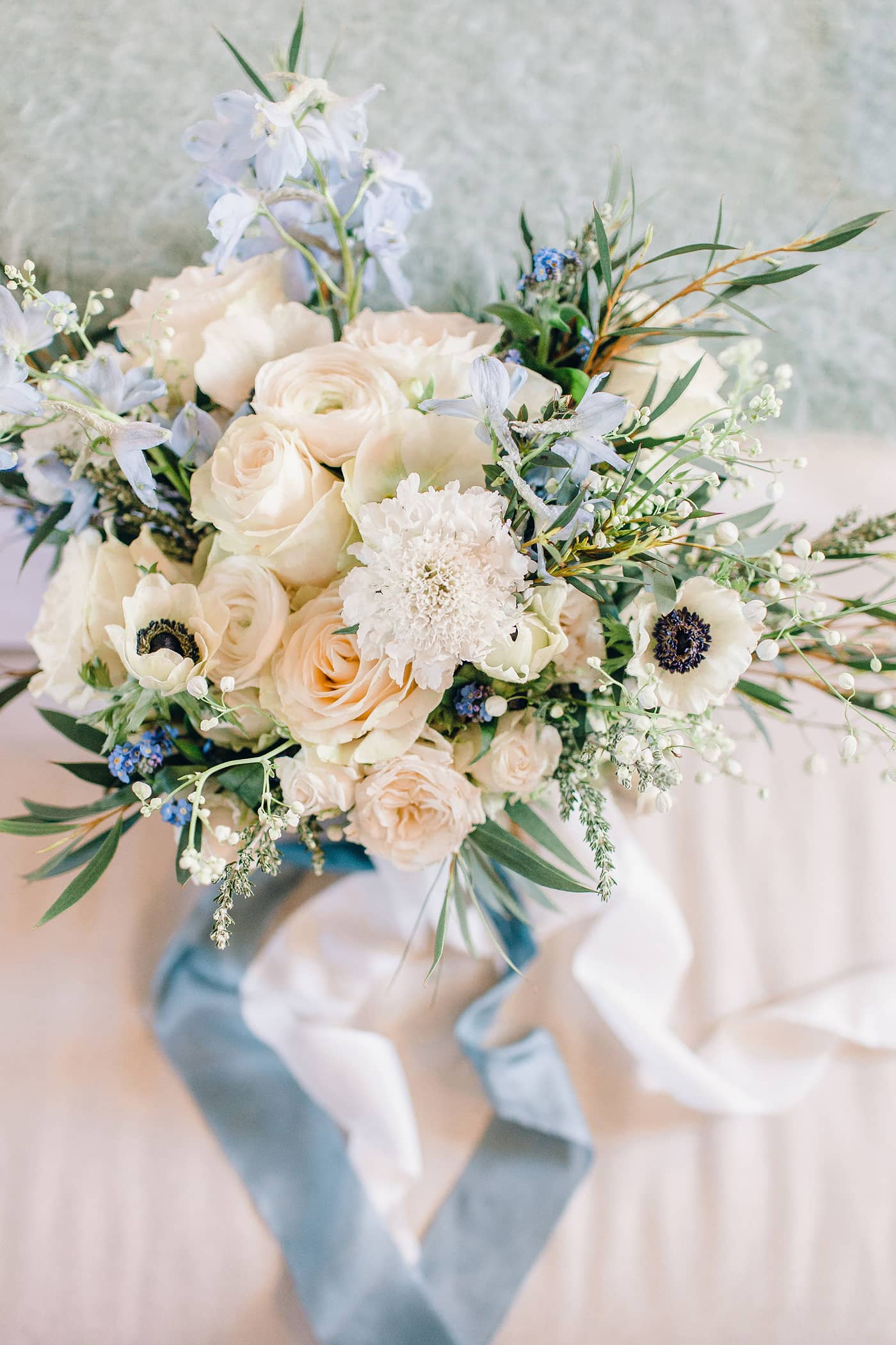 A wedding bouquet of white and blue flowers with trailing blue ribbons sitting on a fabric covered bench at Middleton Lodge, The Fig House.