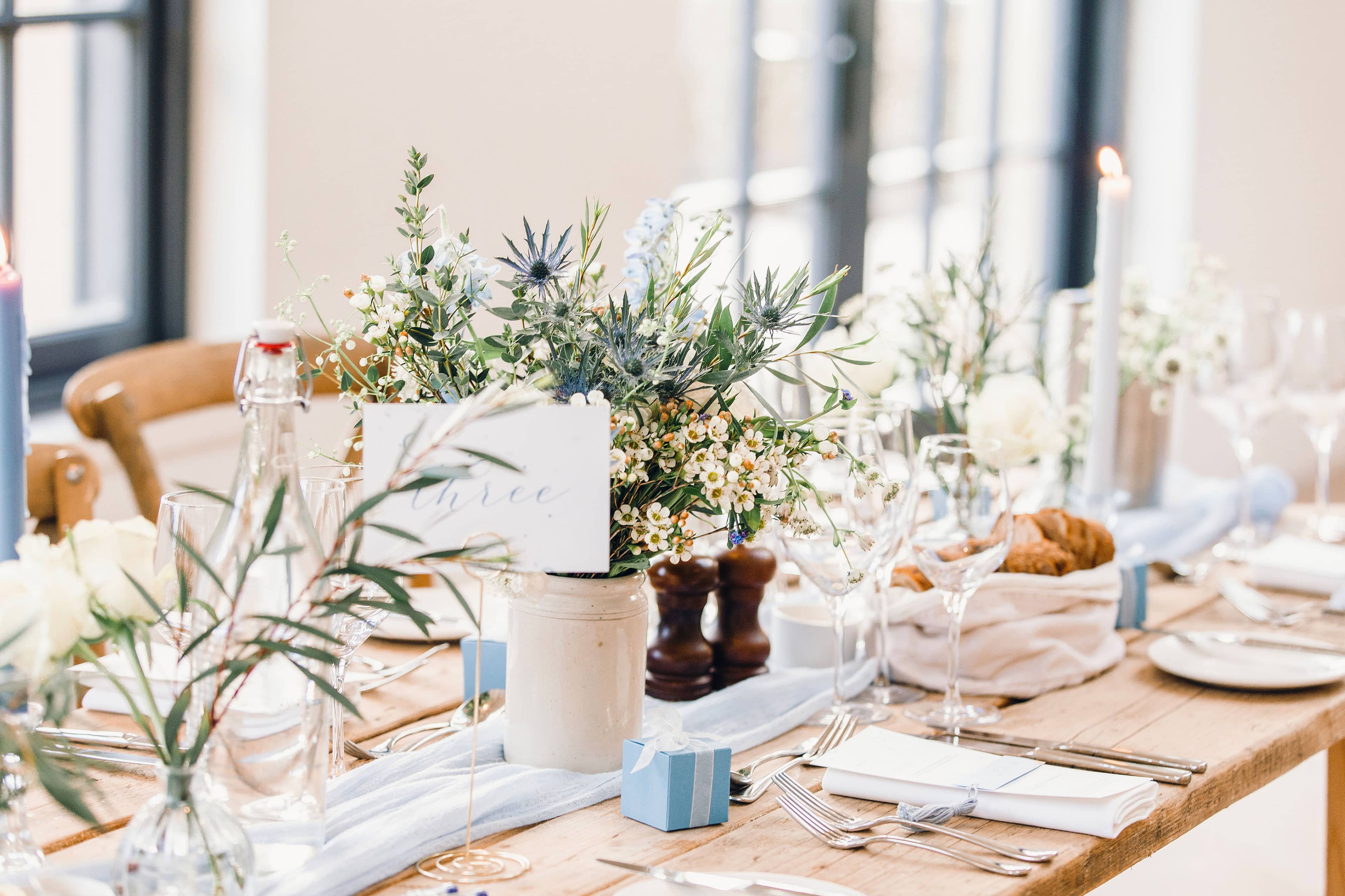 Decorative wedding table setting with white and blue floral arrangements and blue dinner candles in a softly lit room at The Fig House, Middleton Lodge. The tables include blue favour boxes, silver cutlery and bud vases full of white and blue flowers.