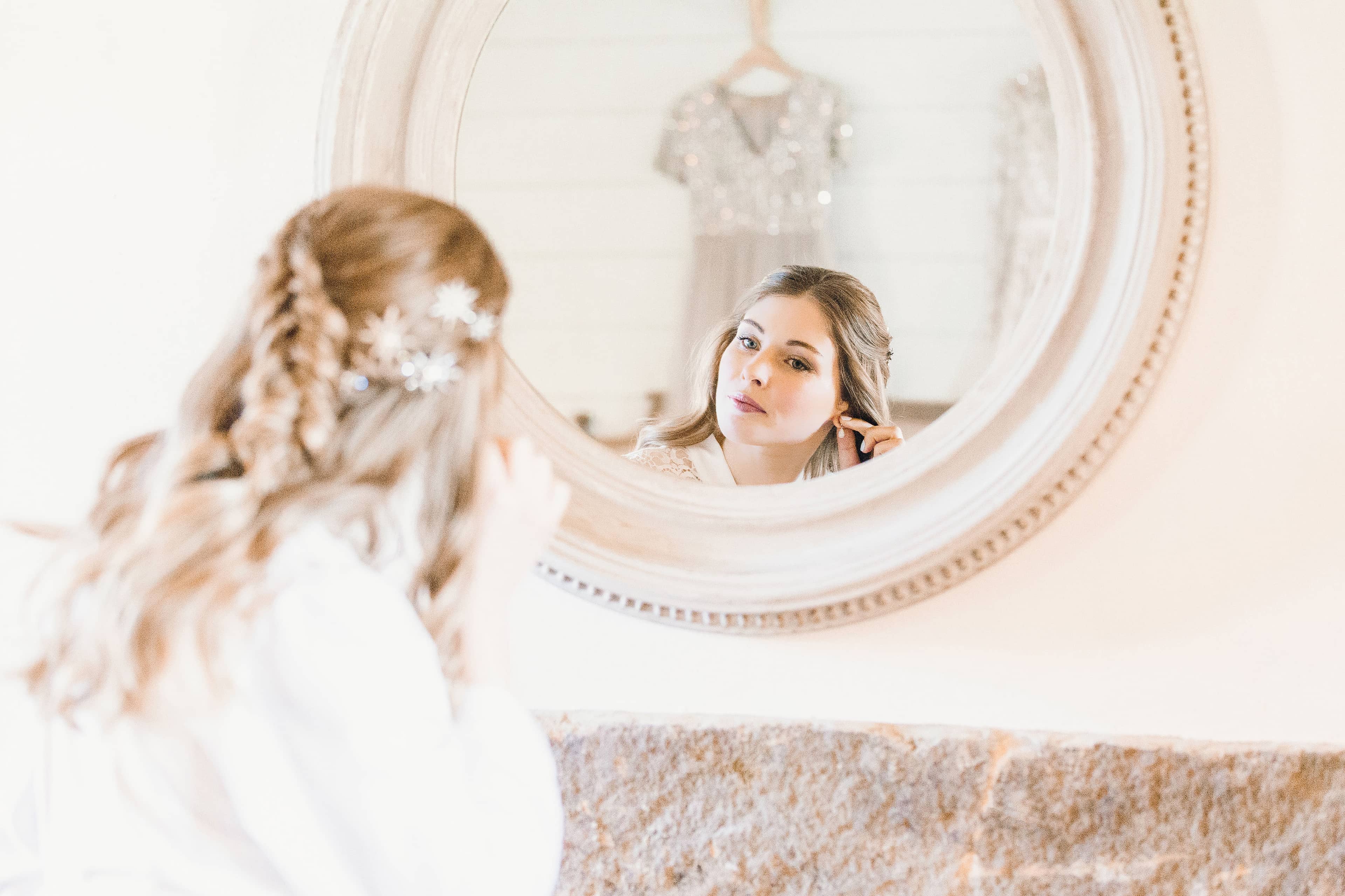 A bride putting in her earrings looking into a round mirror with decorative frame at the bridal suite at The Fig House, Middleton Lodge. There is a reflection of a bridesmaid dress in the mirror.