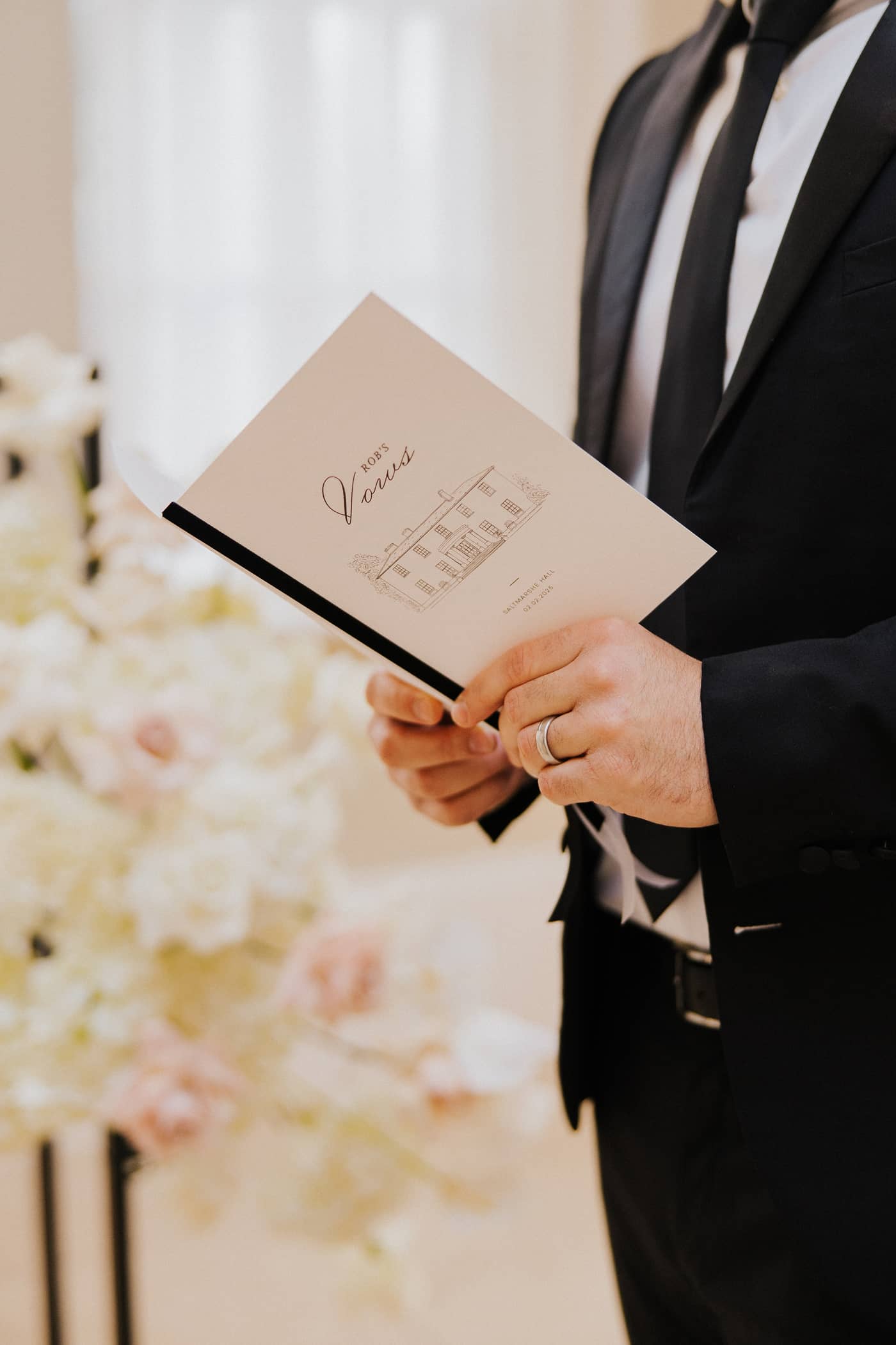 A groom in a black suit holding a wedding vow book, tied with a black velvet ribbon and featuring a line illustration of Saltmarshe Hall on the front. The groom is standing in front of a white and blush floral installation.