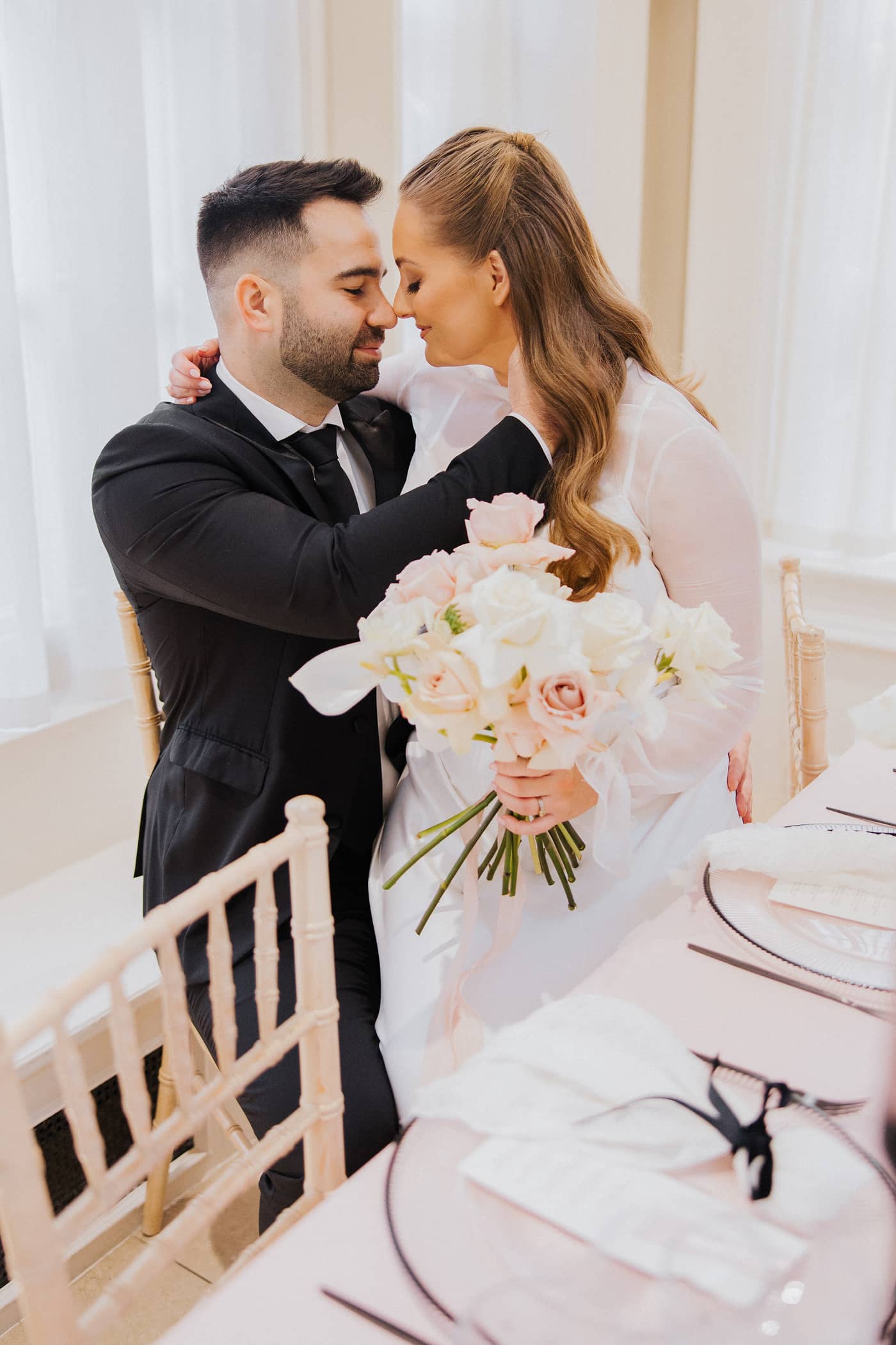 A groom in a black suit and a bride in a silk wedding dress, sitting down at a wedding table at Saltmarshe Hall. The bride is holding a white and blush bridal bouquet and the table is covered in a blush pink tablecloth and decorated with black candlesticks and pink candles.