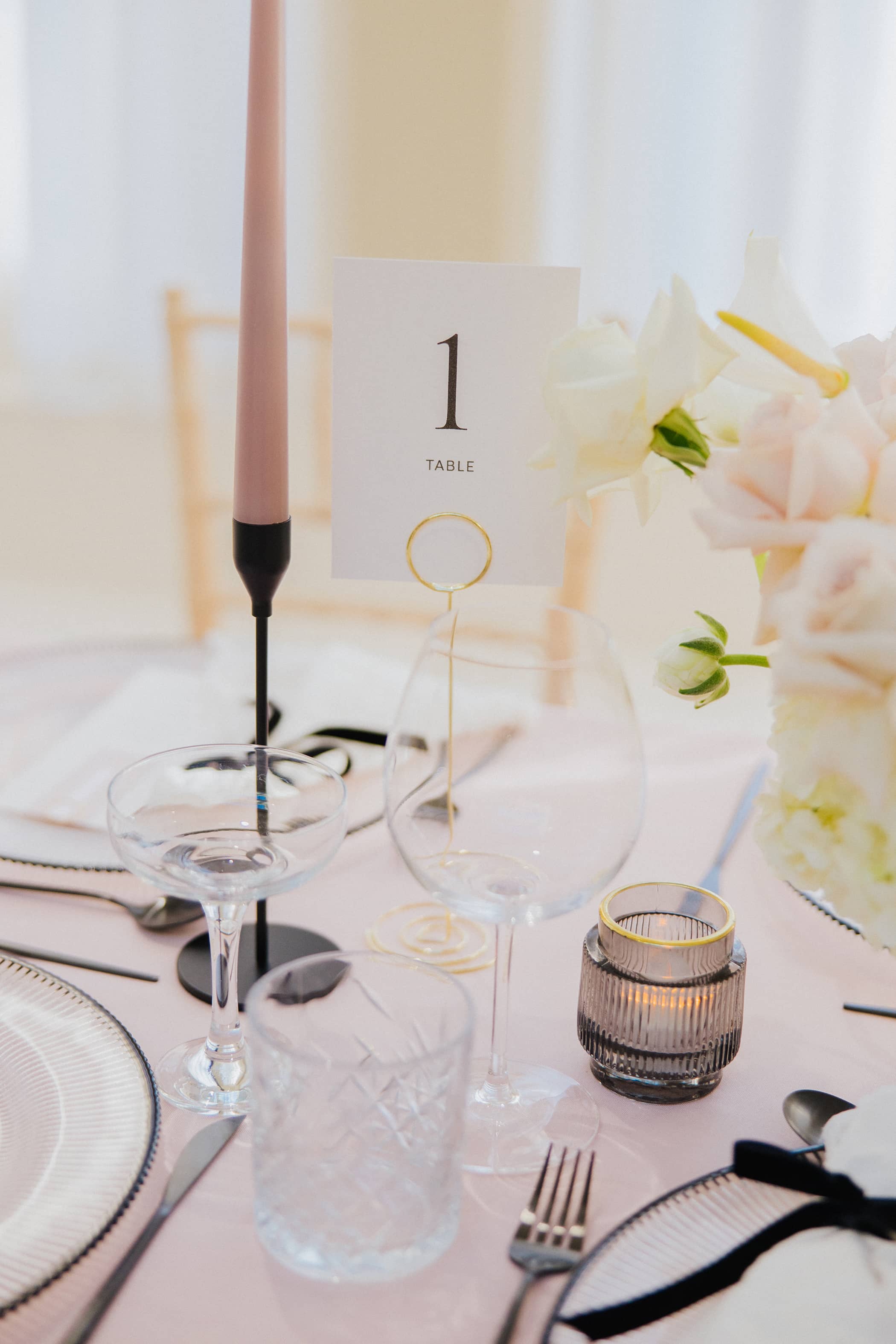 A close up of a table number sign. The wedding table has pastel pink tablecloths, light wooden chairs, black cutlery and candle sticks, pink candles and a white and blush floral arrangement at Saltmarshe Hall.