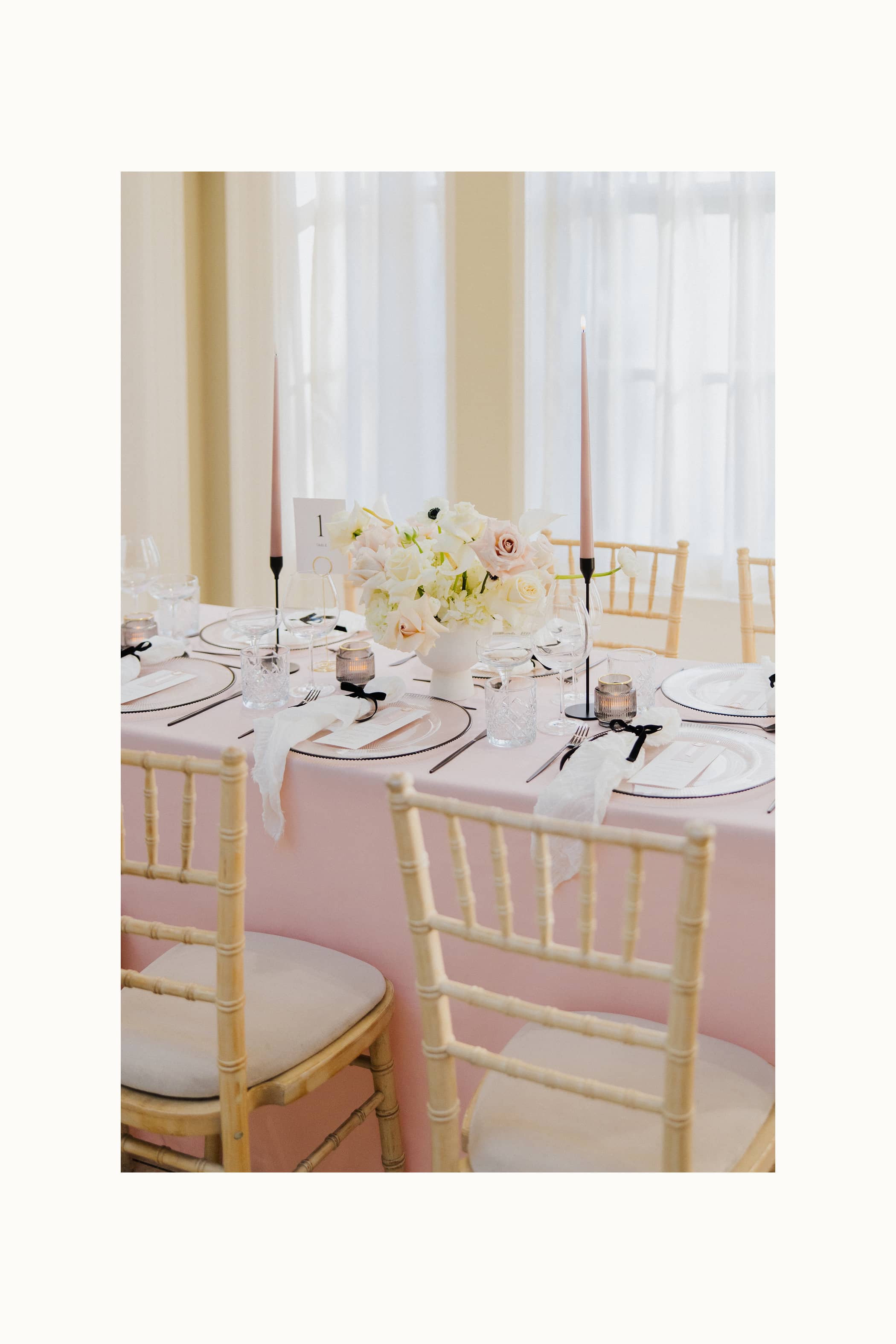 A wedding table with pastel pink tablecloths, light wooden chairs, black cutlery and candle sticks, pink candles and a white and blush floral arrangement at Saltmarshe Hall.
