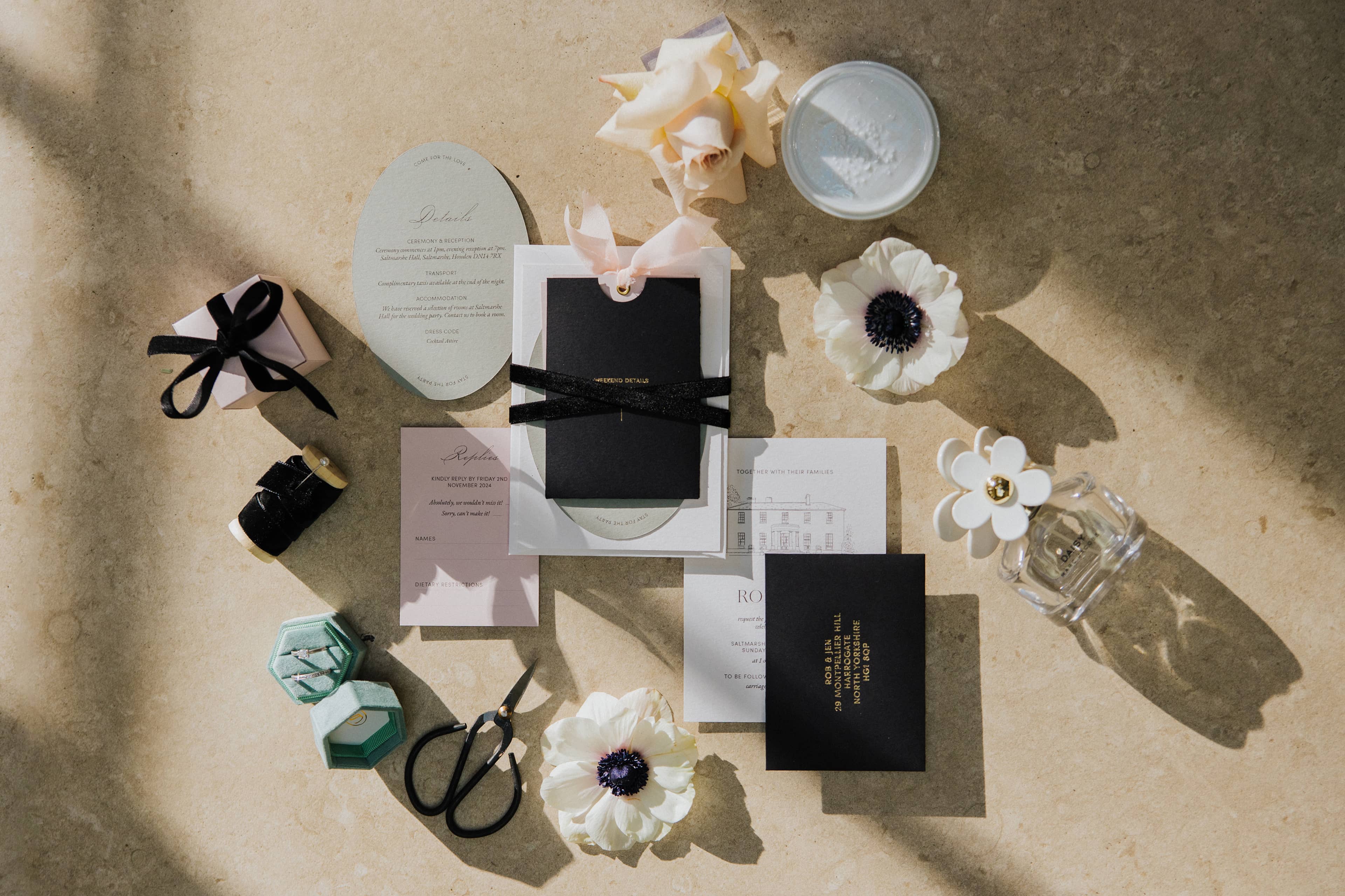 A flatlay of a wedding invitation suite, that features a line illustration of Saltmarshe Hall, gold foil and black and blush pink colours, lying on a textured surface.