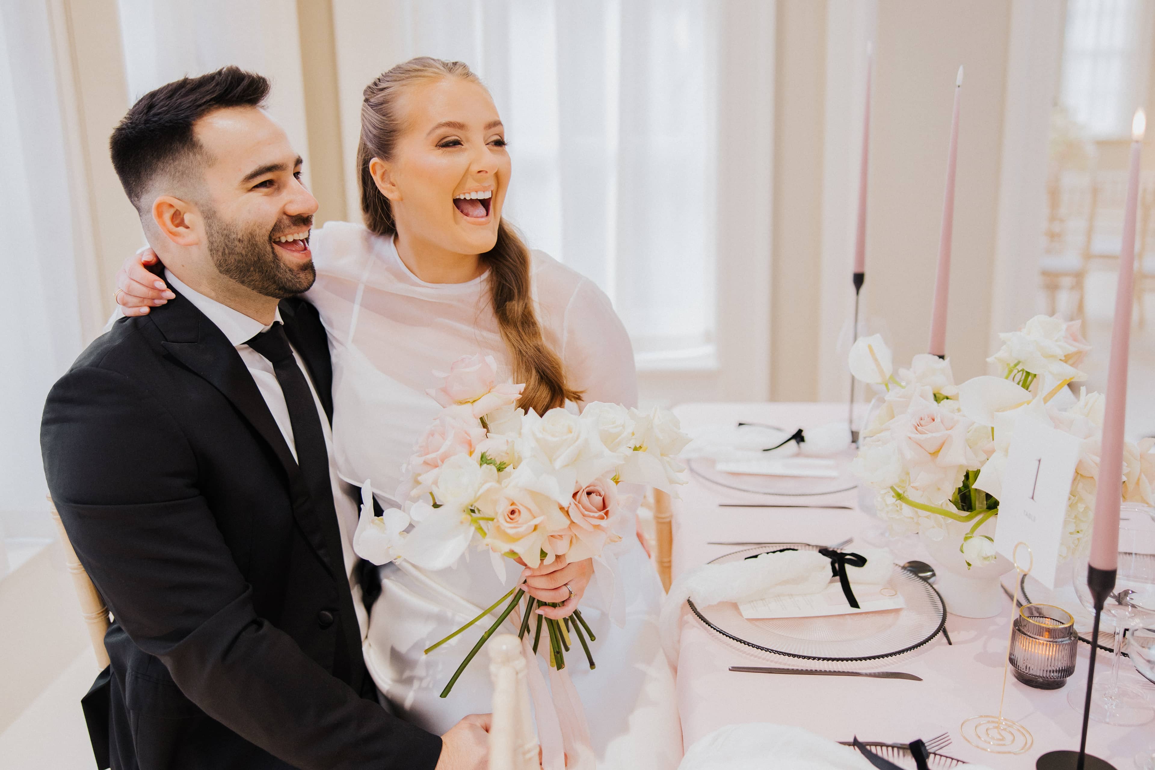 A groom in a black suit and a bride in a silk wedding dress, sitting down and smiling at a wedding table at Saltmarshe Hall. The bride is holding a white and blush bridal bouquet and the table is covered in a blush pink tablecloth and decorated with black candlesticks and pink candles.