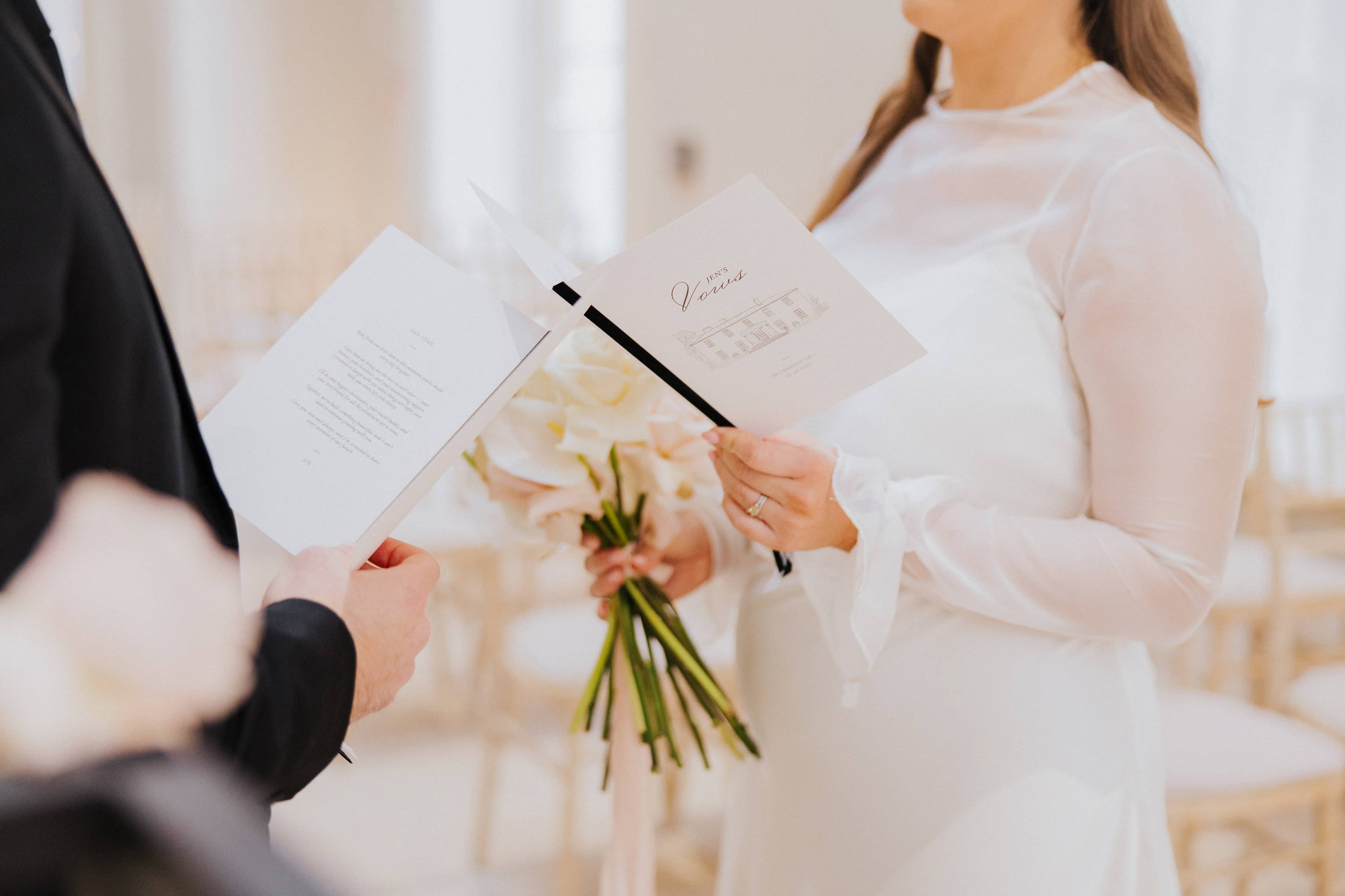A groom in a black suit and a bride in a silk wedding dress, both holding a wedding vow book, tied with a black velvet ribbon and featuring a line illustration of Saltmarshe Hall on the front. They are standing in front of a white and blush floral installation.